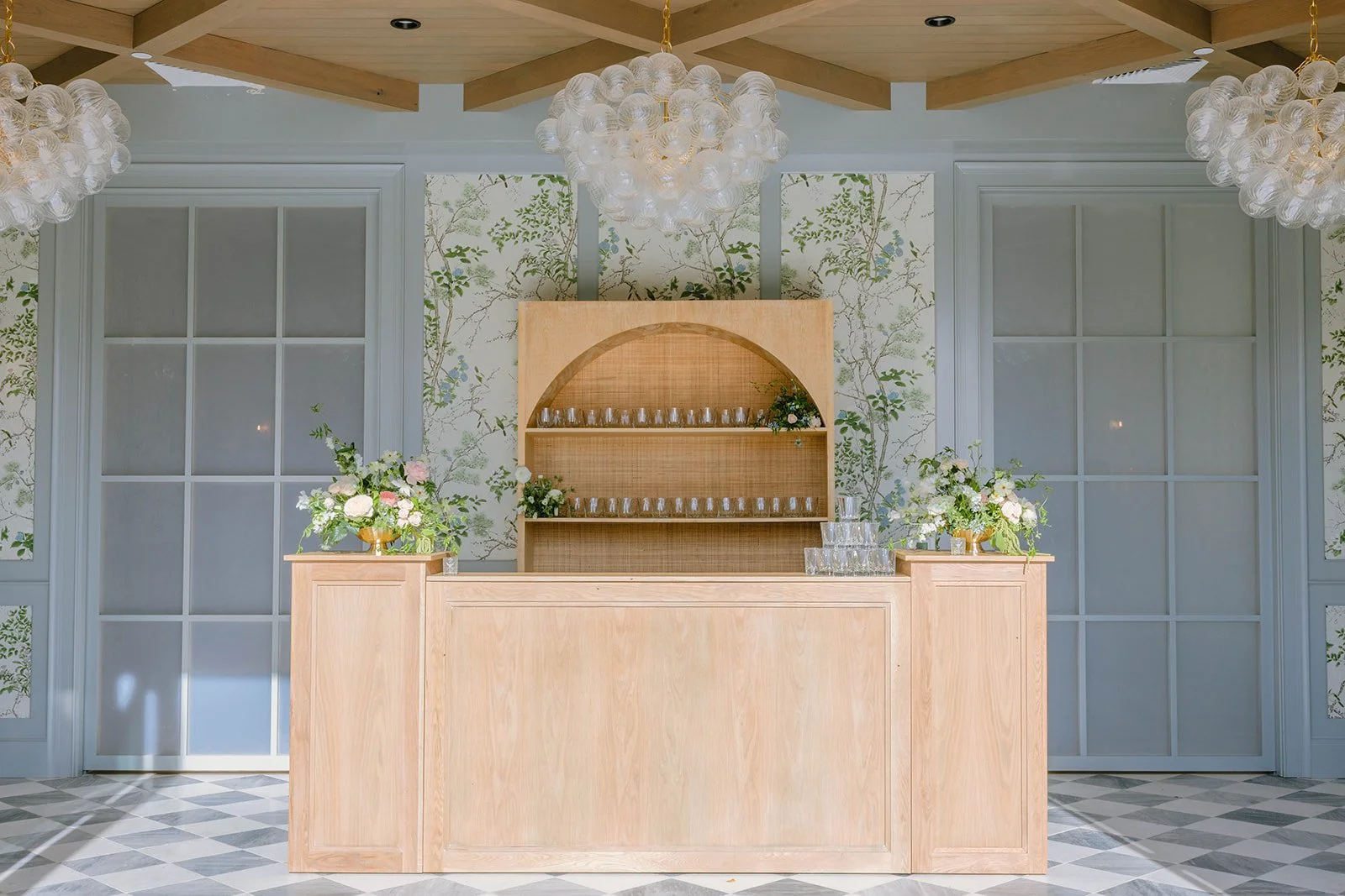 Elegant event bar setup with wooden counter, floral arrangements, and glassware, in a decorated room with patterned floor, floral wallpaper, white panel doors, and hanging chandeliers.