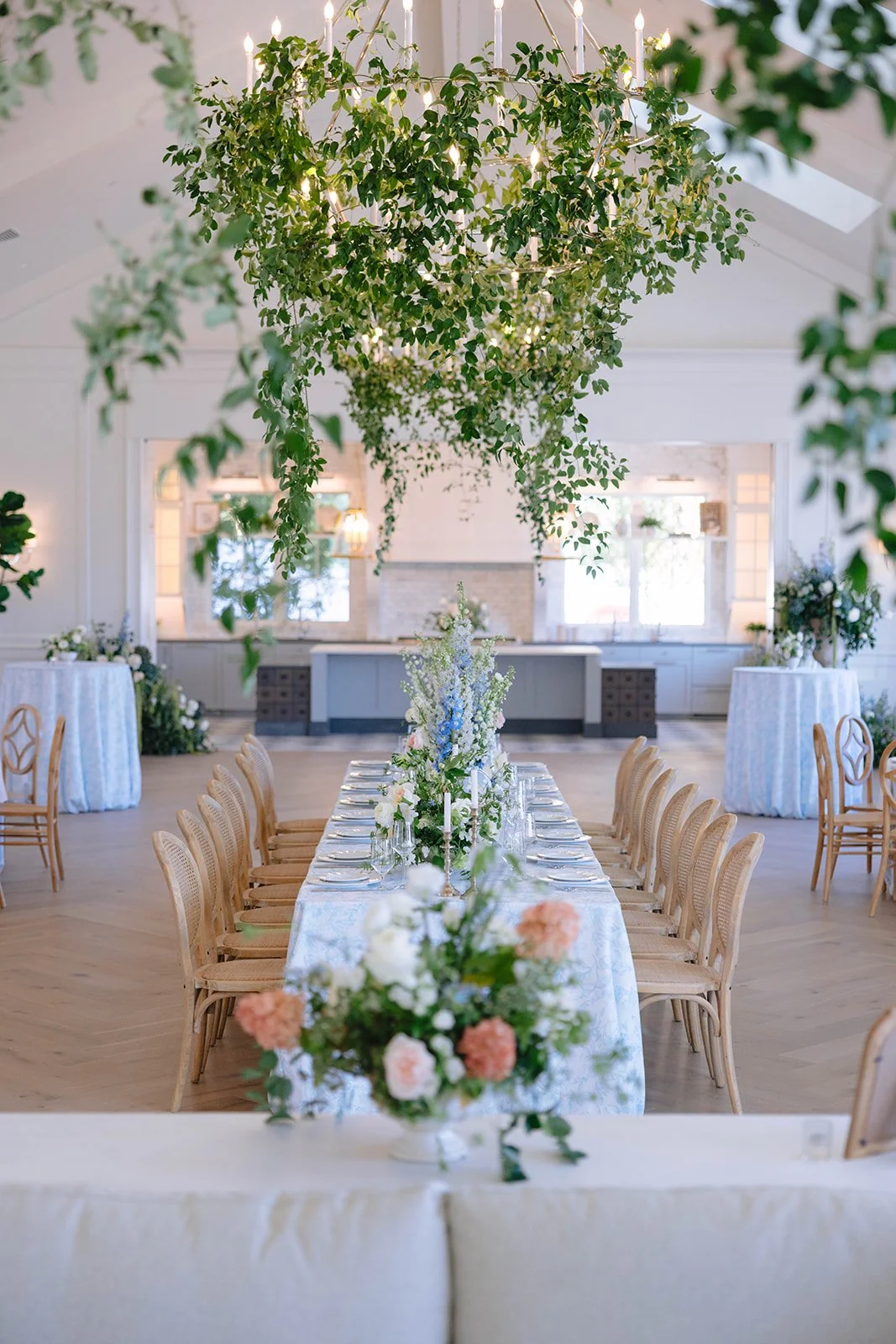 Elegant wedding reception setup with a long table decorated with white and pastel floral arrangements, surrounded by wooden chairs, under a lush hanging chandelier of greenery with candle lights inside, in a bright and airy room with white walls and 