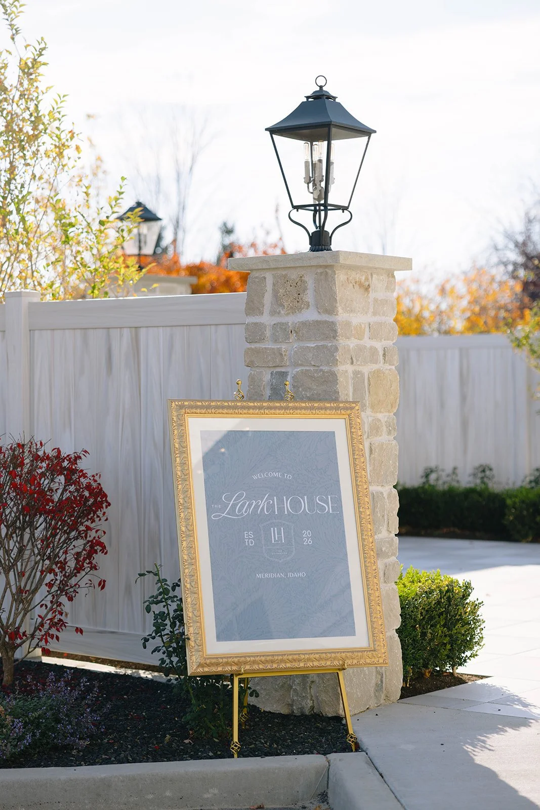 A decorative outdoor sign parades the welcoming message, 'Welcome to The Lark House, Meridian, Idaho, EST 2026,' displayed in a gold frame, set against a stone pillar with a black lantern on top, surrounded by landscaped garden.