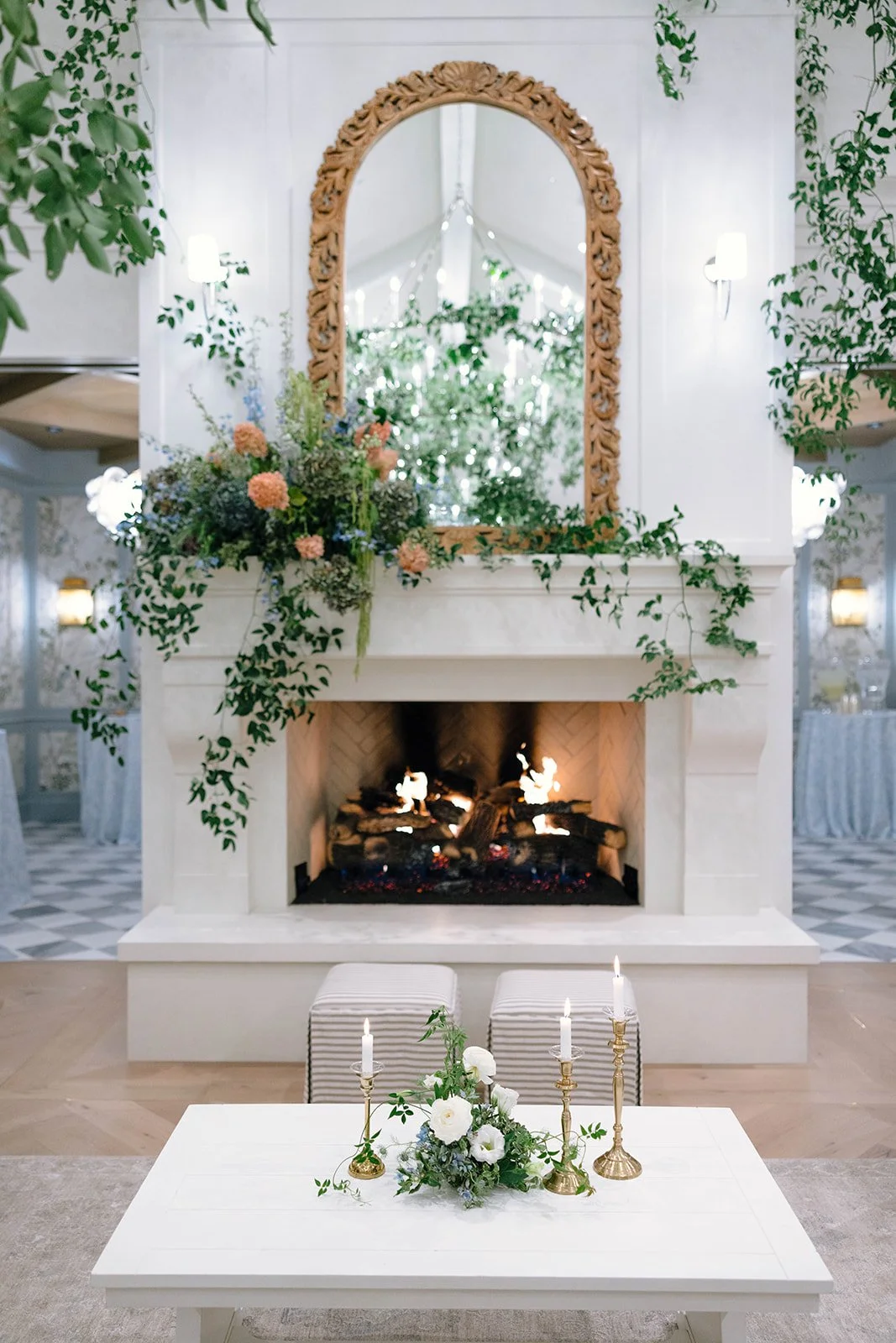 Elegant interior with a fireplace decorated with flowers and greenery, a large mirror above the fireplace, a table with a floral arrangement and candles, and a pristine room in the background.