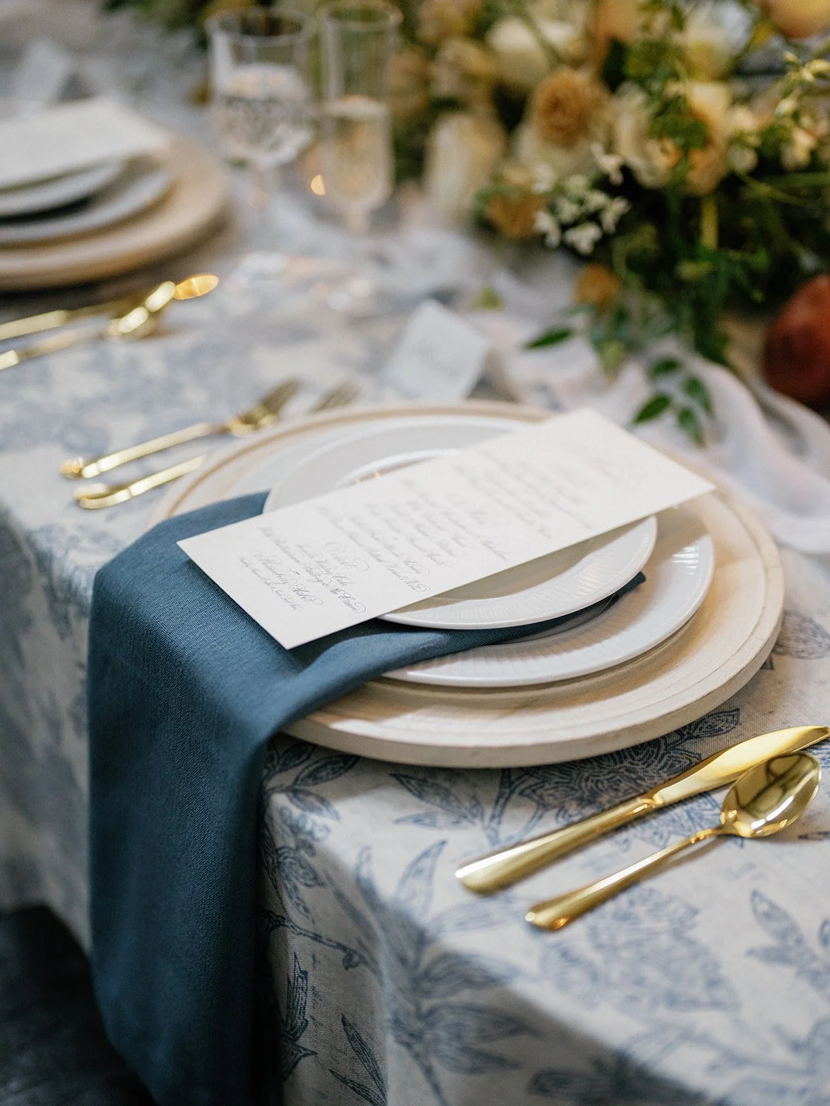 A formal table setting with gold utensils, white plates, a dark blue napkin, a printed menu card, and a floral centerpiece in the background.