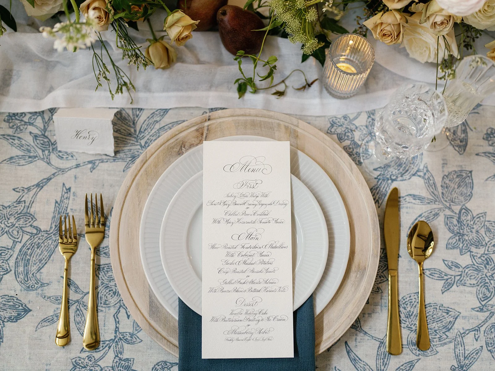 Elegant table setting with a menu card, gold utensils, glassware, candles, and a floral centerpiece.