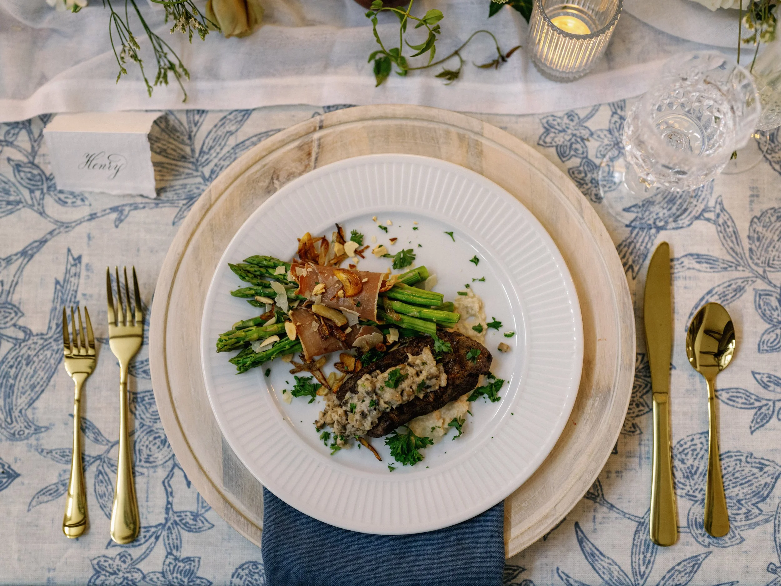 A plated meal on a table with gold utensils, a blue napkin, and a place card labeled "Henry." The dish includes asparagus, toasted almonds, and a meat entrée topped with a stuffing or sauce.