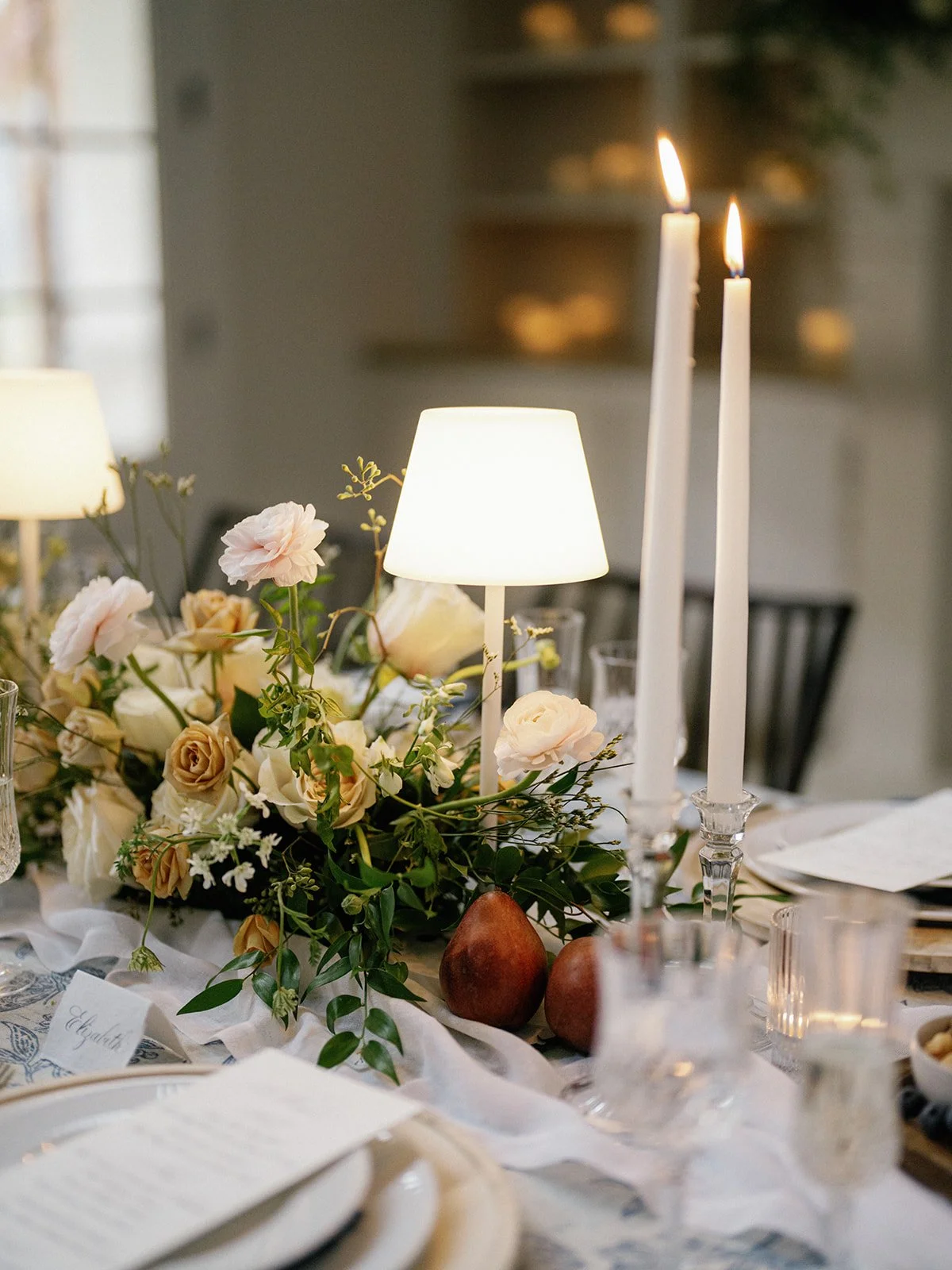 A decorated dining table featuring a floral centerpiece with cream and blush roses, two lit tall white candles in glass holders, a small white lamp, and other tableware and decor items.