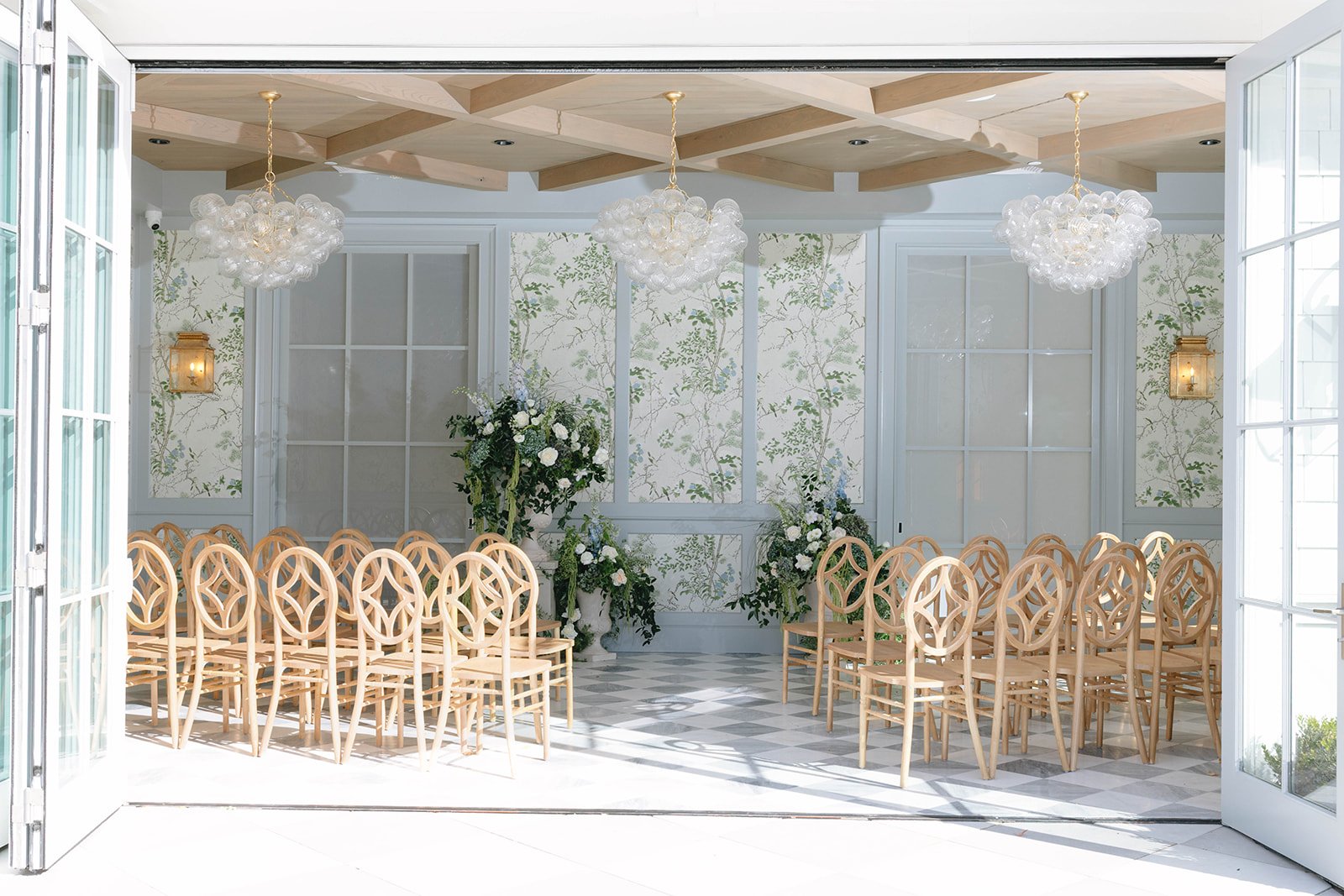 An elegant indoor wedding ceremony setup with wooden chairs arranged in rows, floral arrangements at the front, and modern light fixtures hanging from the ceiling. Double glass doors open to a sunlit outdoor space.