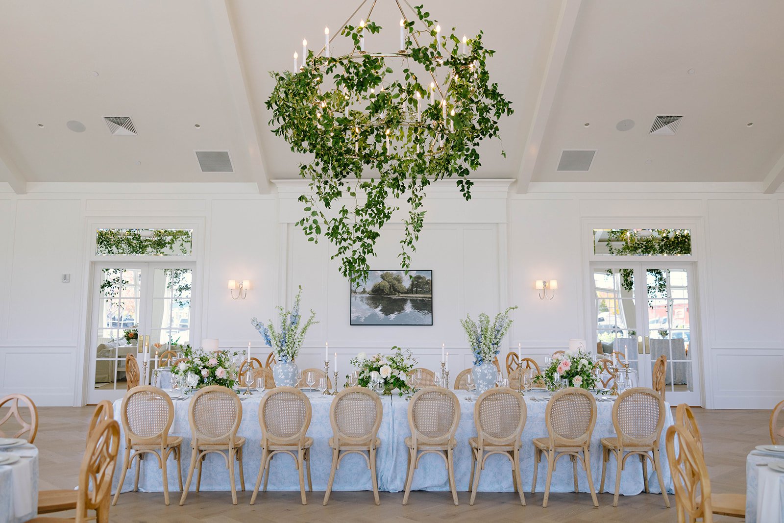 Elegant banquet room decorated for a celebration with a long table adorned with white tablecloth, floral centerpieces, and tall candelabras. Greenery chandelier hanging from the ceiling and framed landscape painting on the white wall. Large windows w