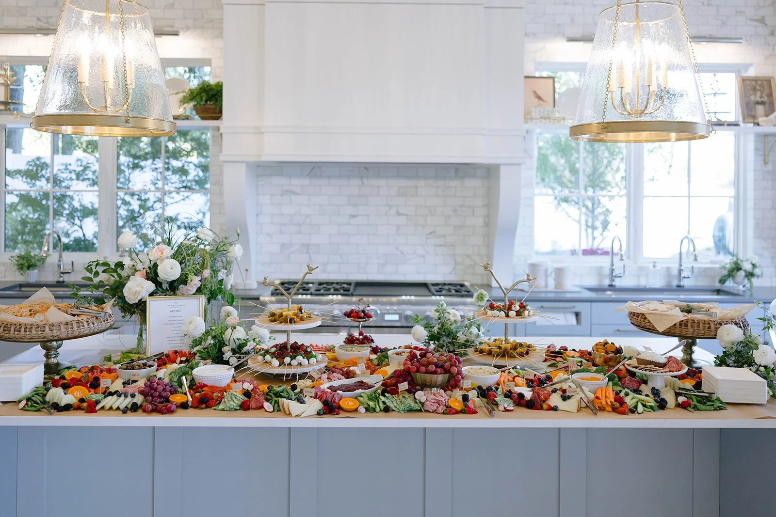A kitchen counter decorated as a food spread with fruit, cheese, and snacks, featuring floral arrangements, under large hanging lamps, with white brick walls and windows in the background.