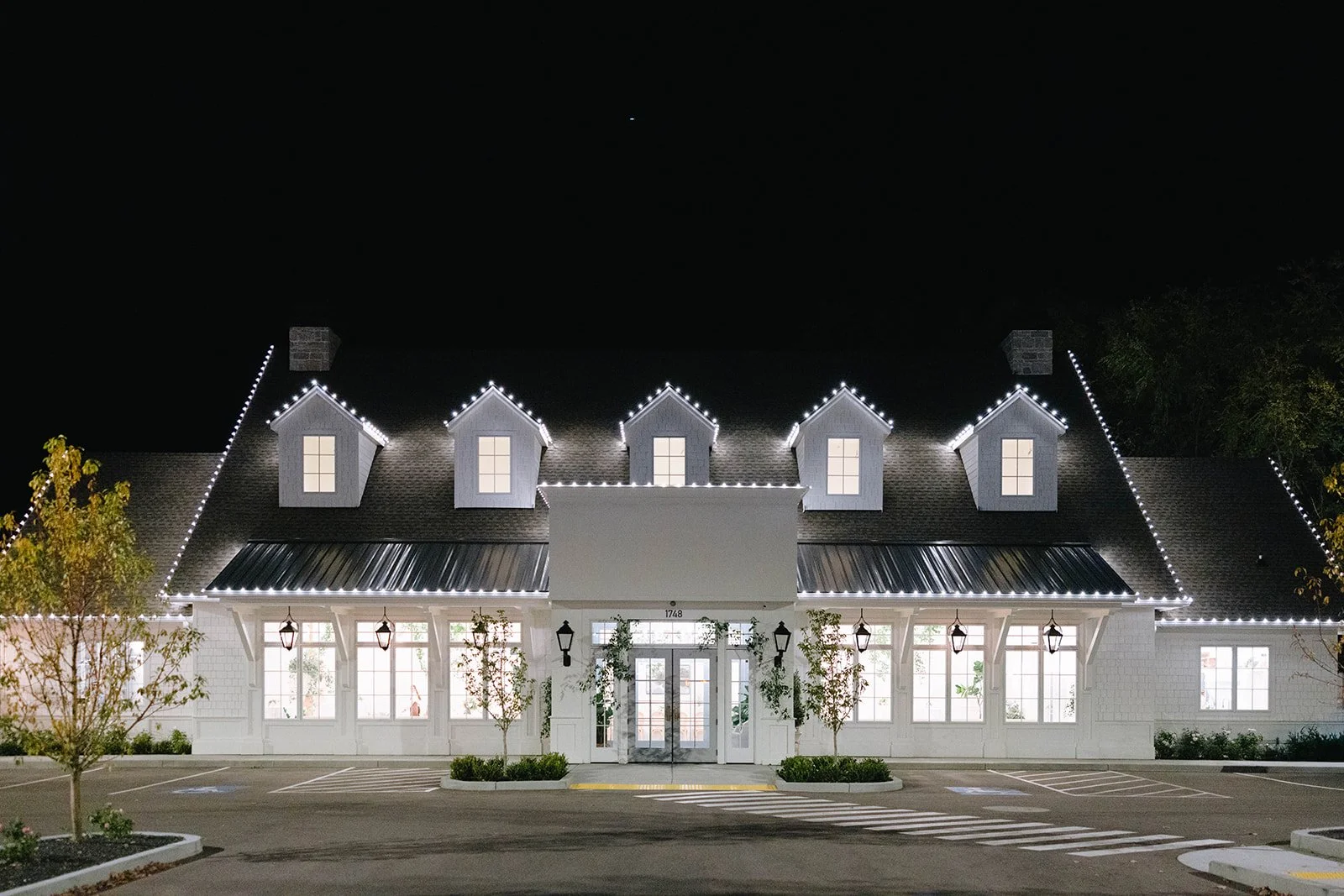 Exterior of a white building at night decorated with string lights on roof edges and dormer windows, with a parking lot in front and trees on the sides.
