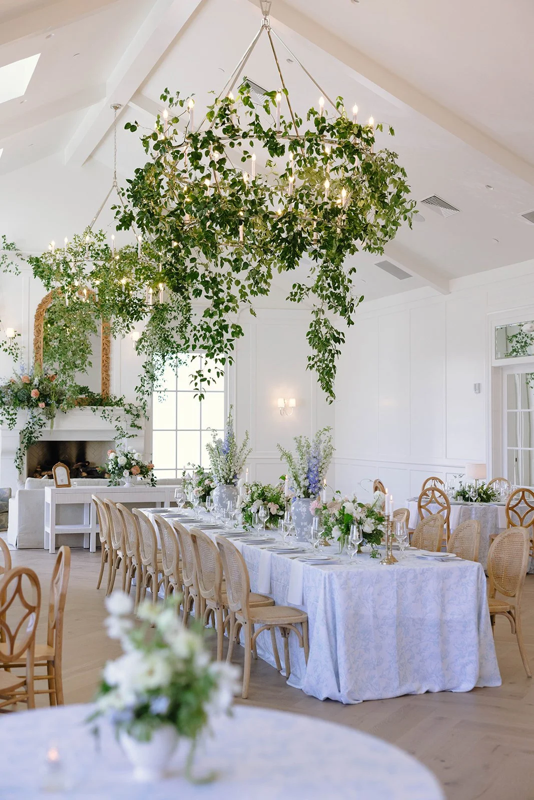 A decorated dining area with white tablecloths, floral centerpieces, and hanging greenery from the ceiling, set in an elegant, bright room with white walls and large windows.