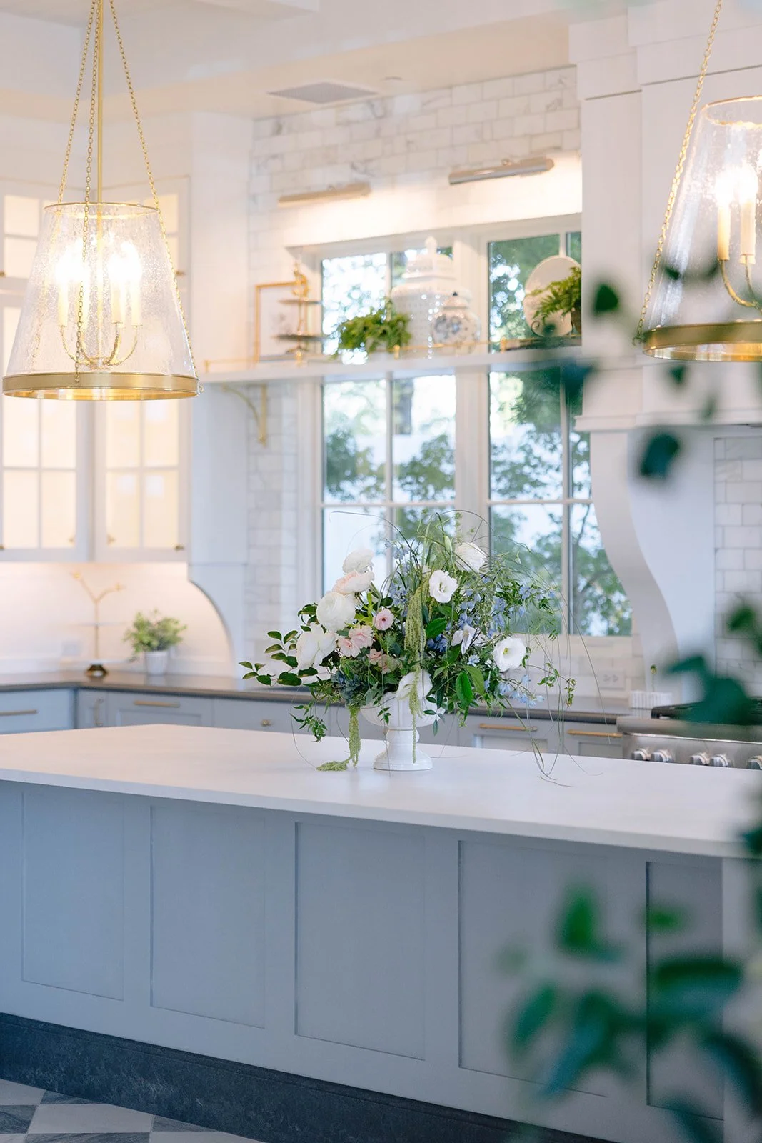 Bright kitchen with white cabinetry, a white countertop island, hanging pendant lights, large windows, leafy green plants, and a floral arrangement in a white vase on the island.