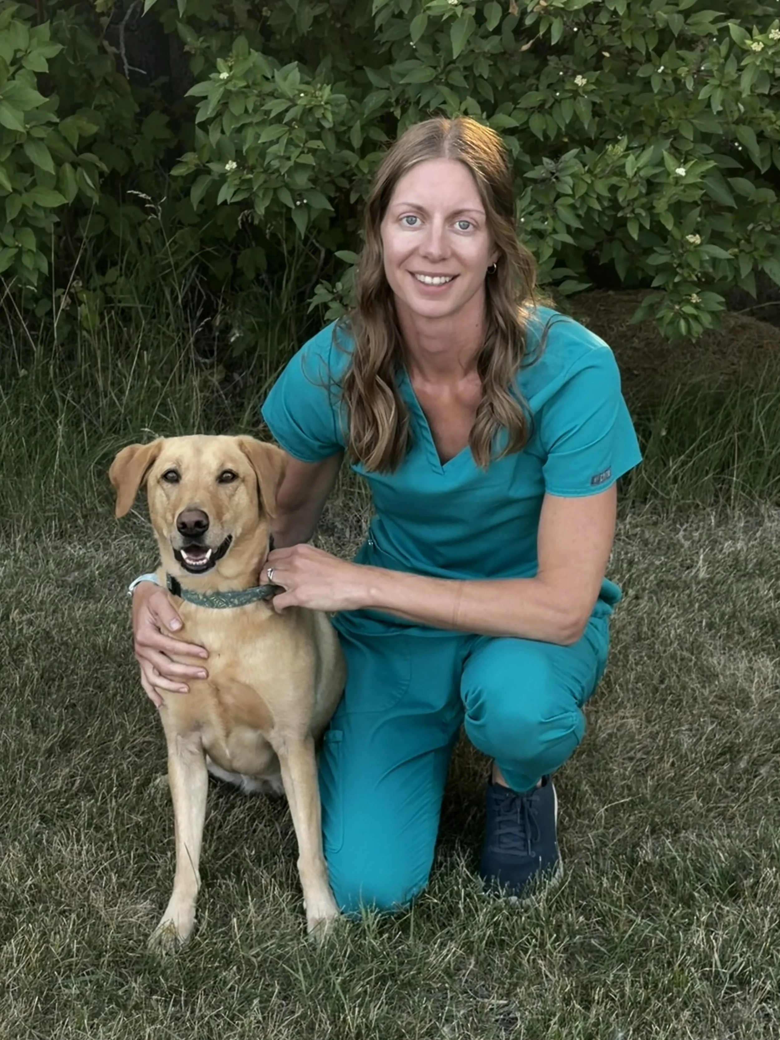 A woman with long, light-brown hair is kneeling in a lawn with her arm affectionately around a yellow lab sitting to her right.