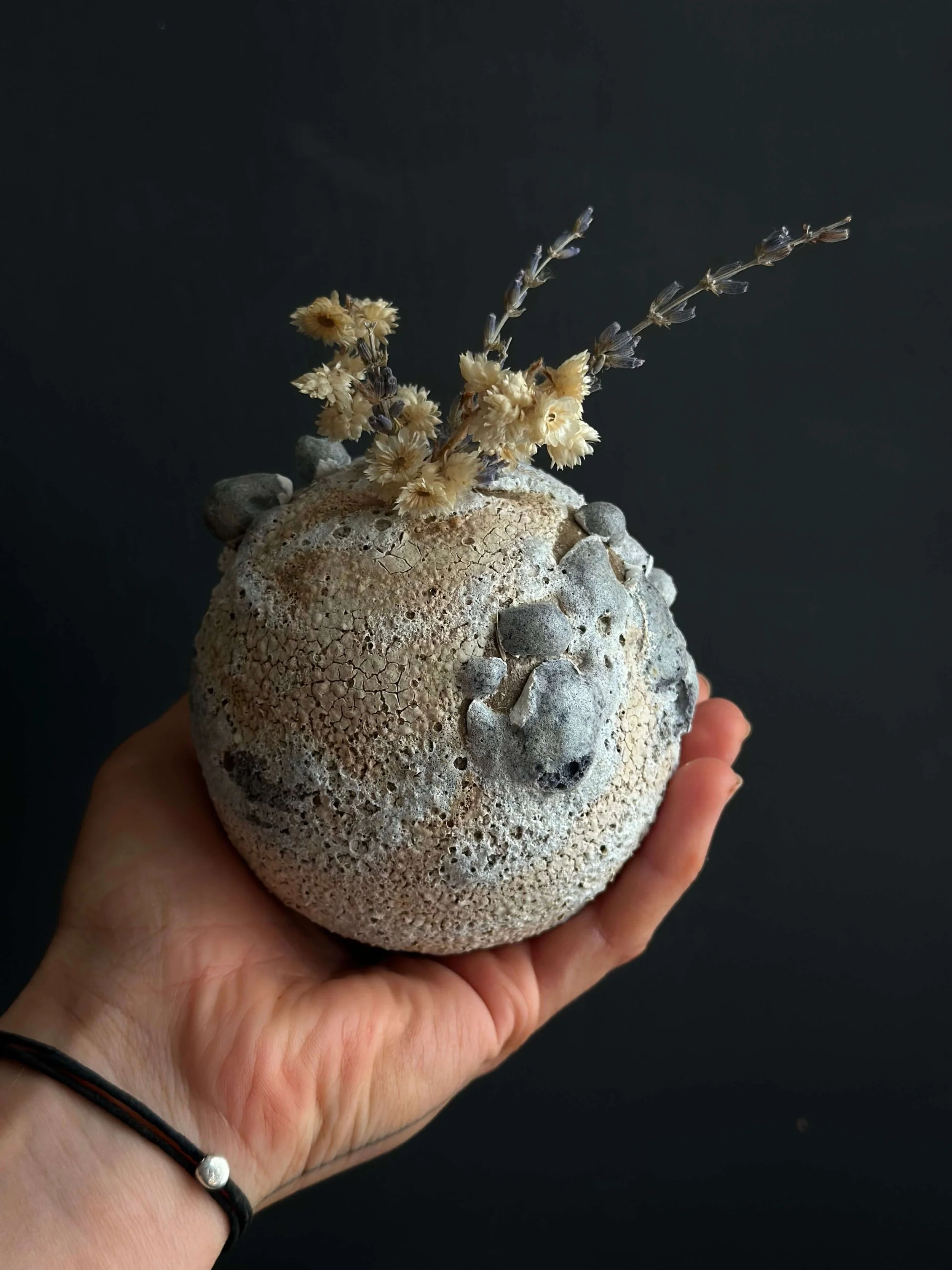 Hand holding small round lava-glazed vessel with dried botanicals