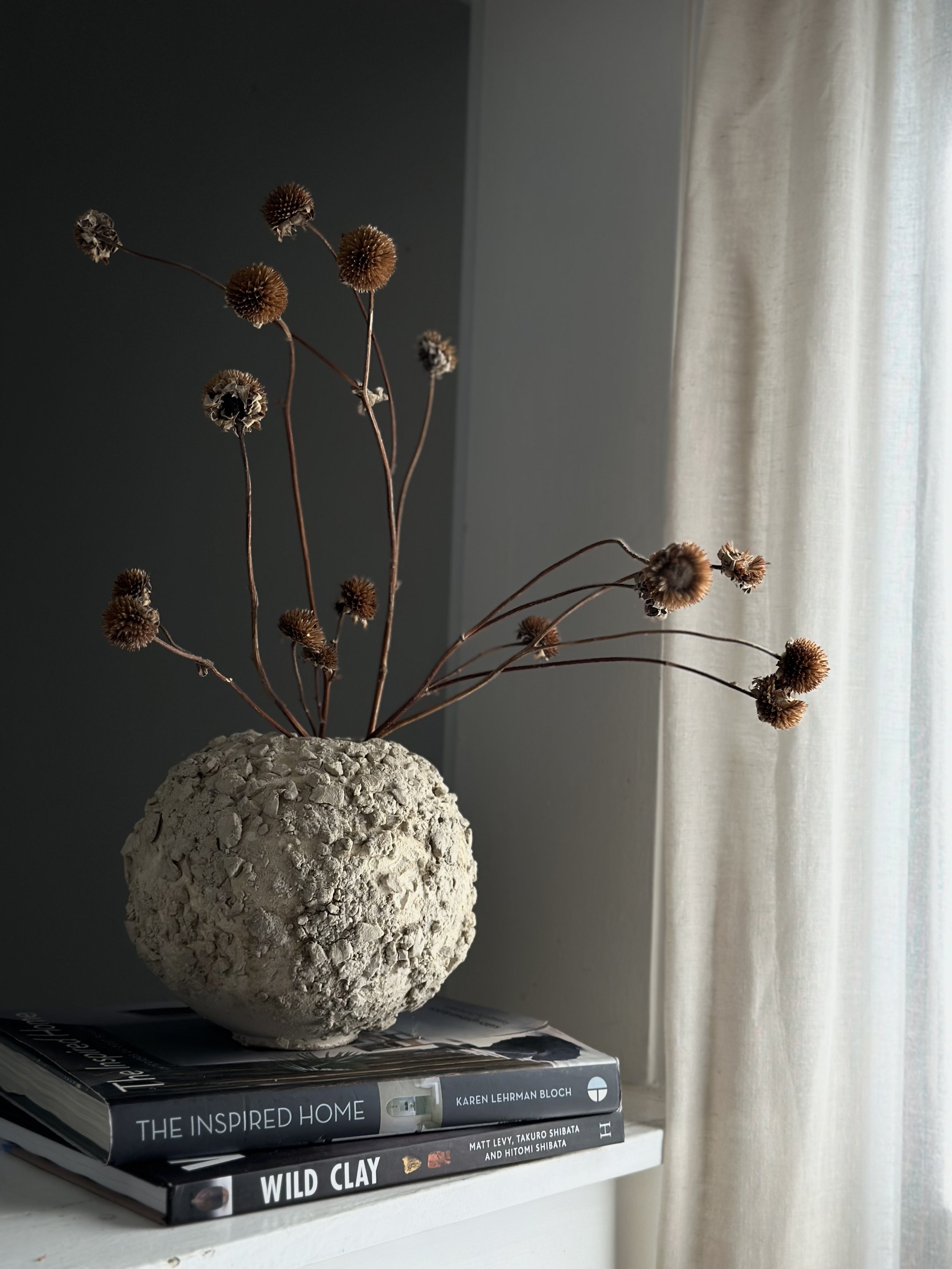 Stone-like ceramic vessel with dried wildflowers on design books, linen curtain