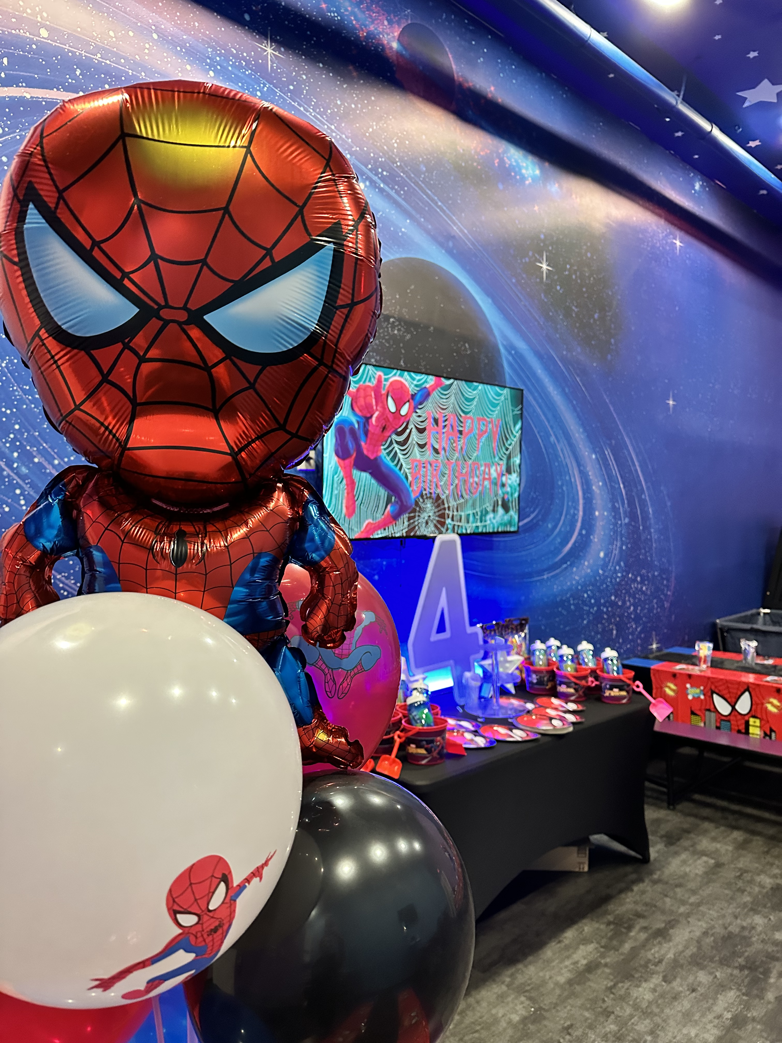 Party decorations with Spider-Man theme, including balloons and a 'Happy Birthday' sign in the background, with a space-themed backdrop.
