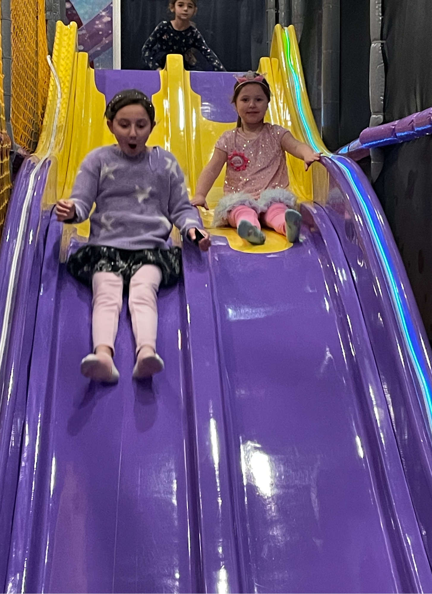 Three young girls playing on a colorful indoor slide at a trampoline park. Two girls are sliding down the purple slide, while one girl is at the top, preparing to slide down.