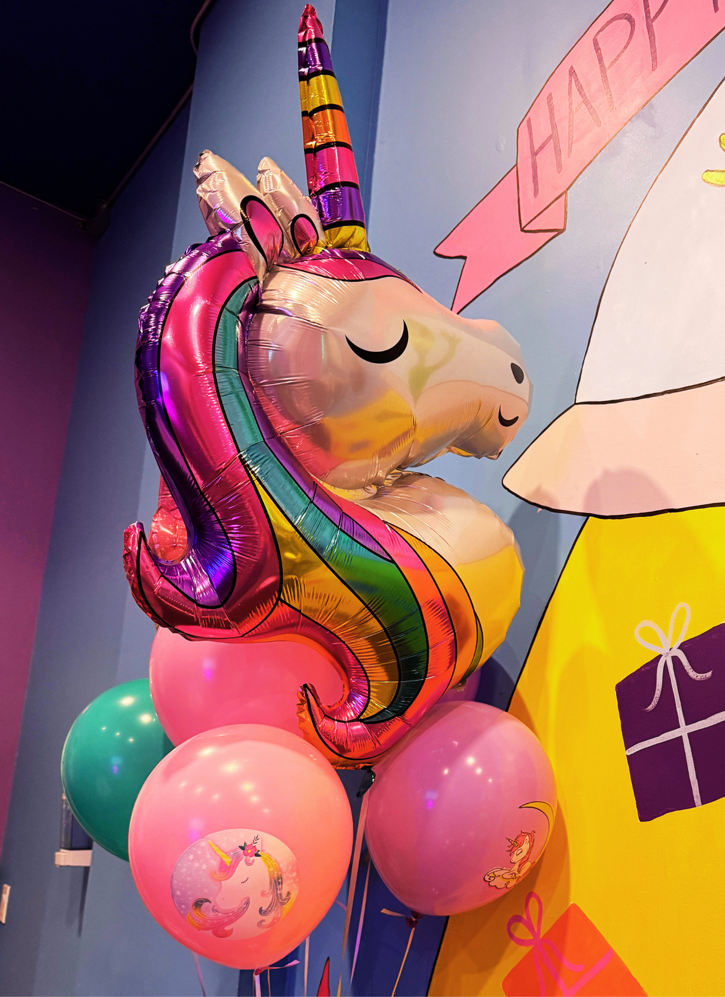 Colorful birthday party decorations including a large unicorn-shaped balloon with rainbow colors, pink and purple balloons with unicorn illustrations, and a pink banner with the word "HAPPY" on a blue wall.