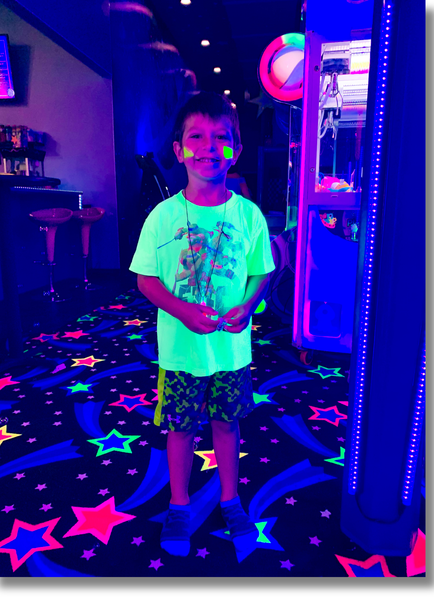 A boy smiling inside a neon-lit arcade with glow-in-the-dark stars on the carpet and illuminated game machines in the background.