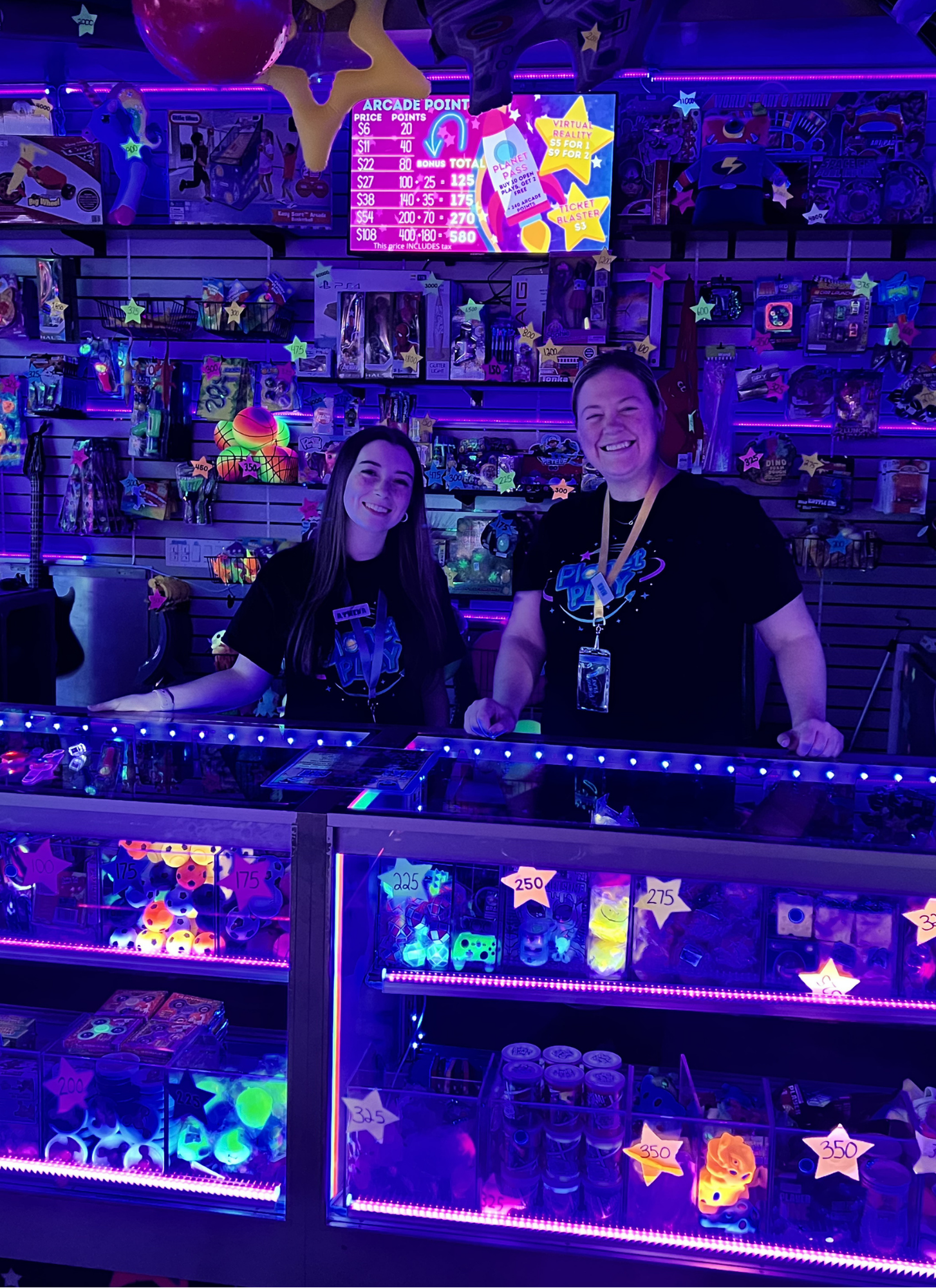 Two women standing behind a brightly lit arcade prize counter filled with toys and prizes in a game arcade. The background features a purple and pink neon glow with a digital score chart and various toys on display.