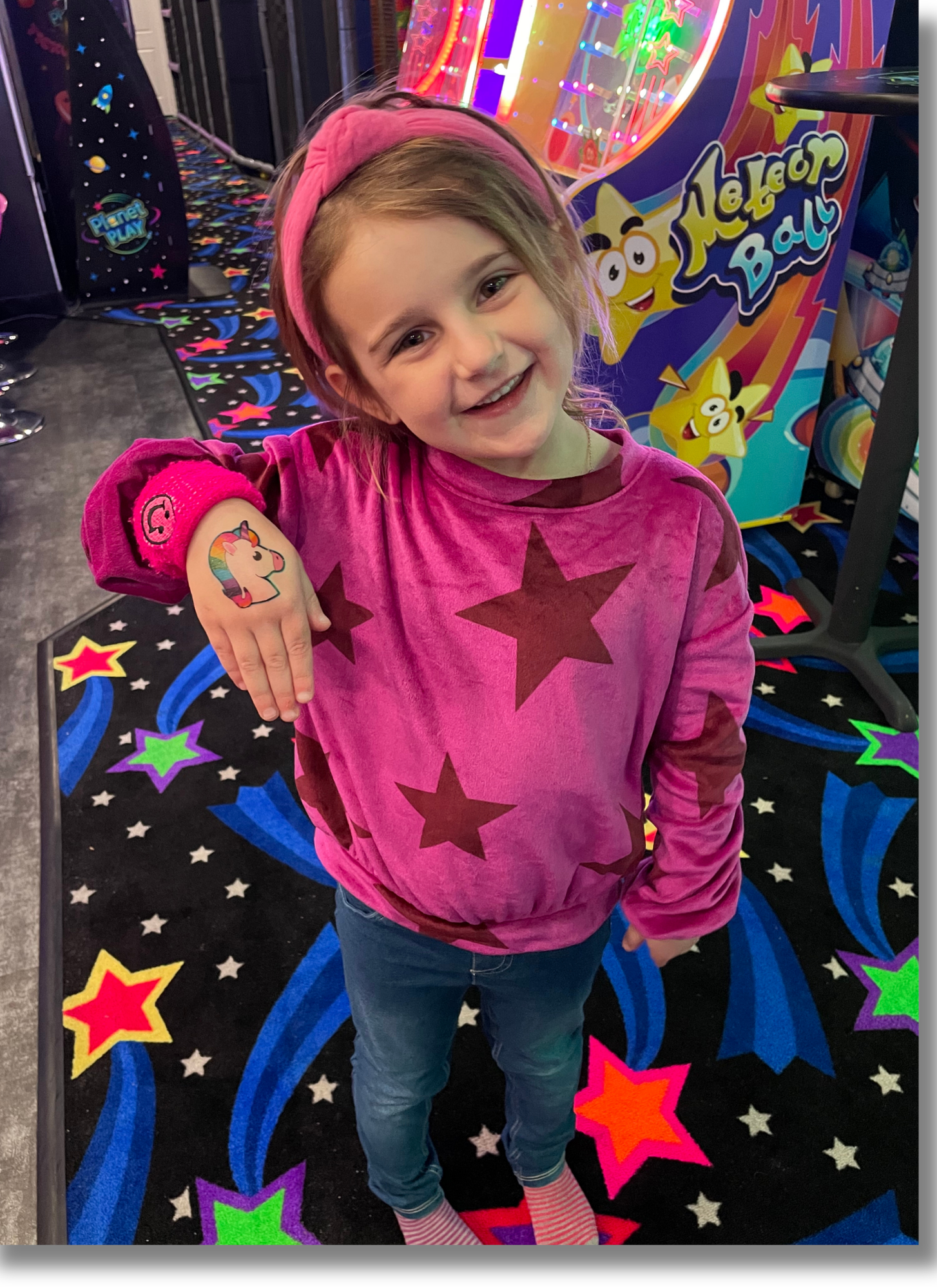 A young girl smiling and showing her rainbow unicorn temporary tattoo on her left forearm at an arcade or game center with colorful star and swirl carpet design and a space-themed game machine in the background.