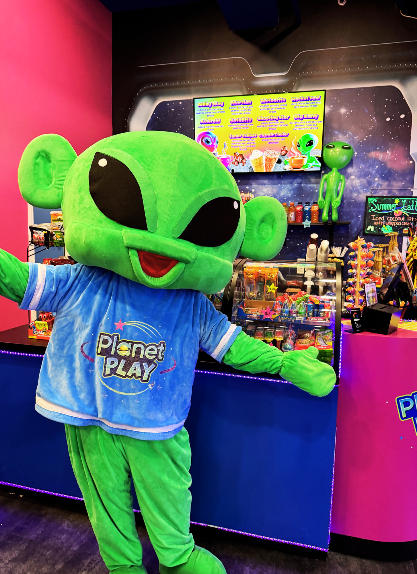Bright green alien mascot with large black eyes and a big smile standing in front of a colorful display at an amusement or themed entertainment venue. The mascot is wearing a blue shirt with "Planet Play" written on it, and the background features a digital menu and snack shelves with colorful candies and drinks.