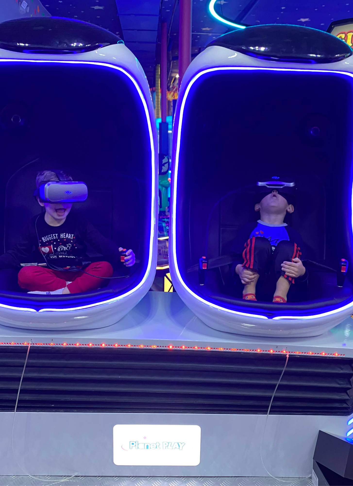 Two children sitting in virtual reality simulator pods at Planet Play, wearing VR headsets, in an arcade setting with blue neon lighting.