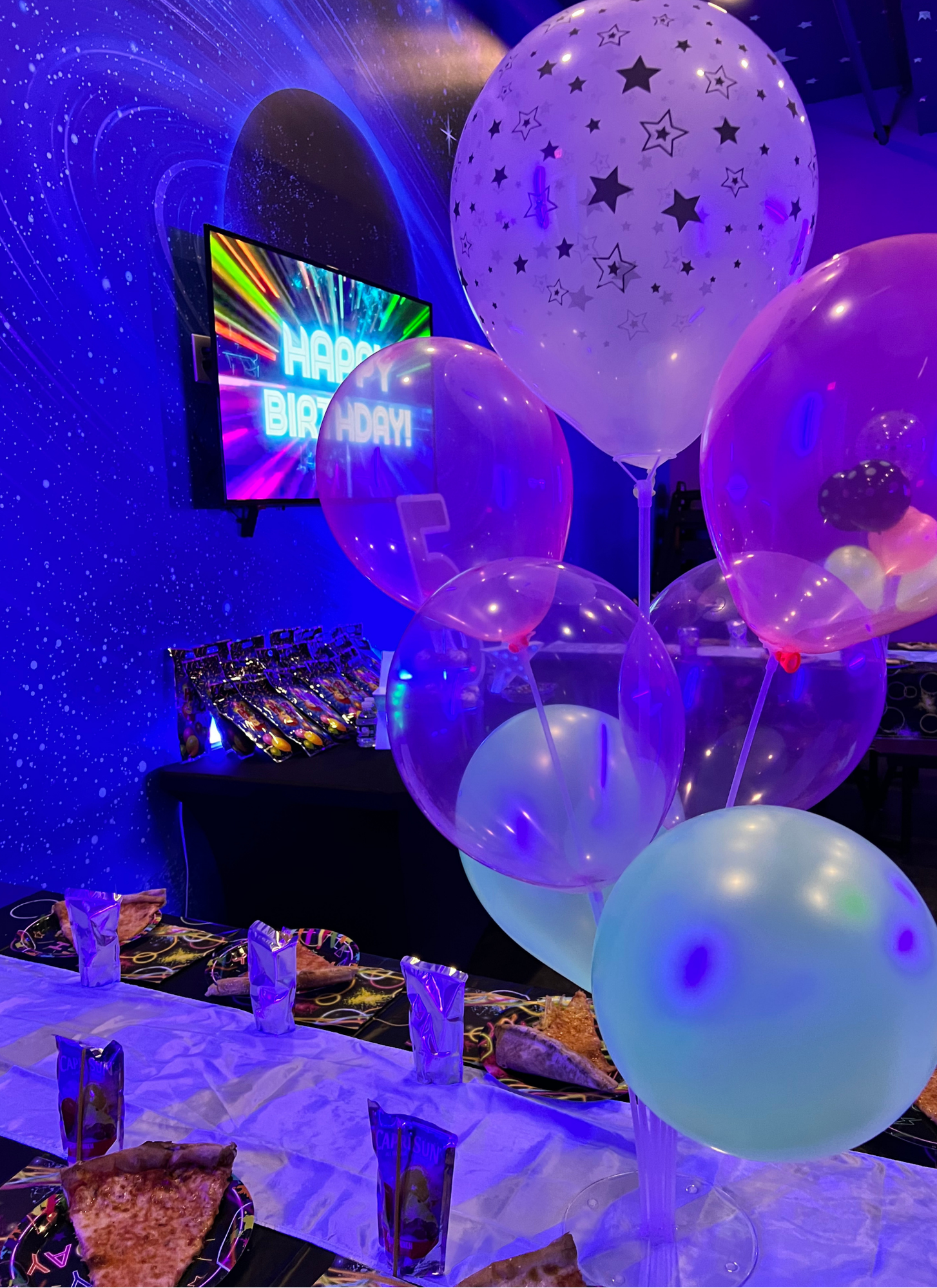 Colorful birthday party scene with balloons, a table with pizza slices, and a TV screen displaying a "Happy Birthday" message with vibrant colors.