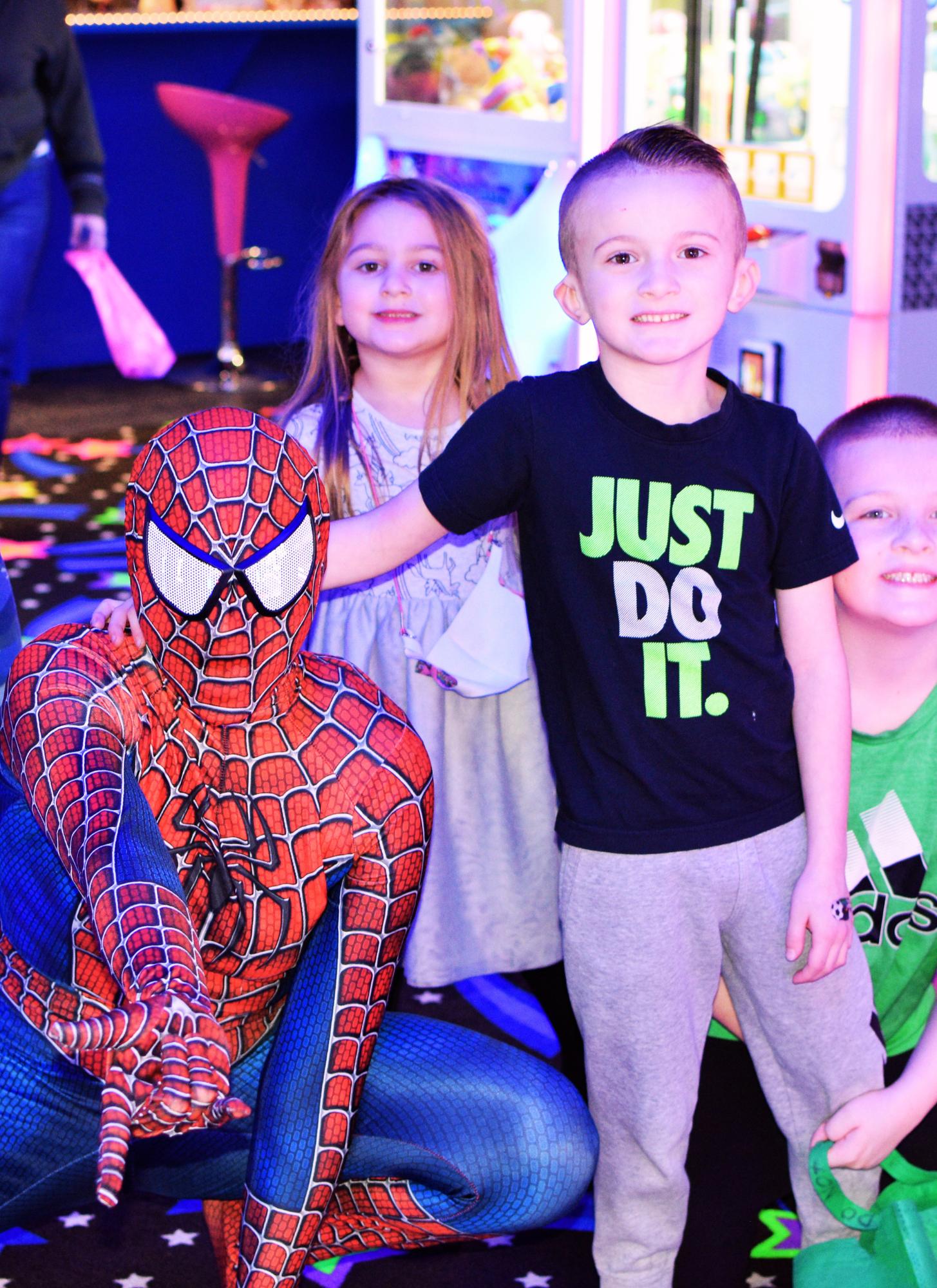 Children at an arcade, one dressed as Spider-Man, and others smiling, with arcade games and toys in the background.