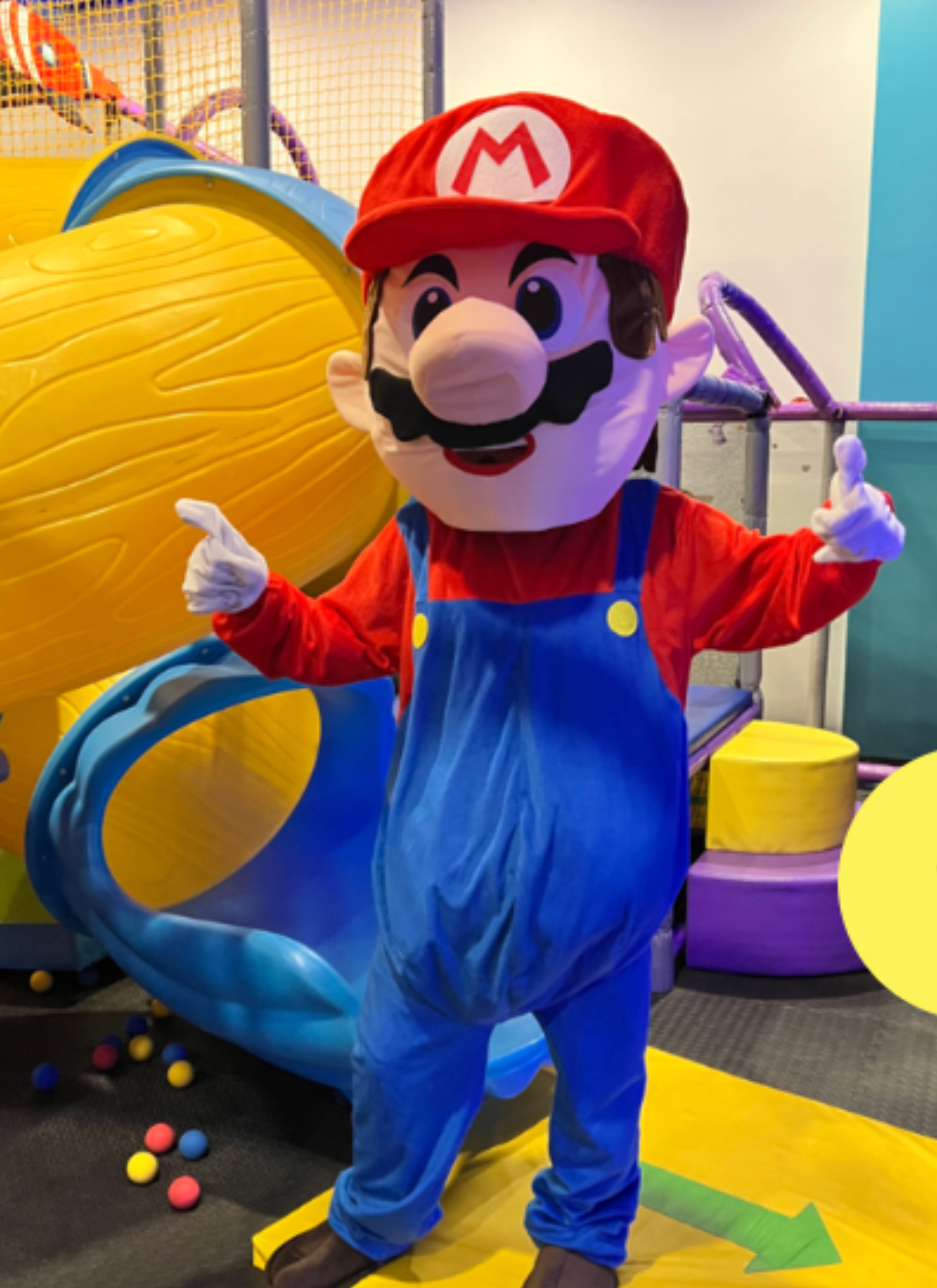 Person dressed as Mario from Super Mario, wearing a red cap with an 'M' emblem, red shirt, blue overalls, and a fake mustache, standing indoors with colorful play equipment and balls around.