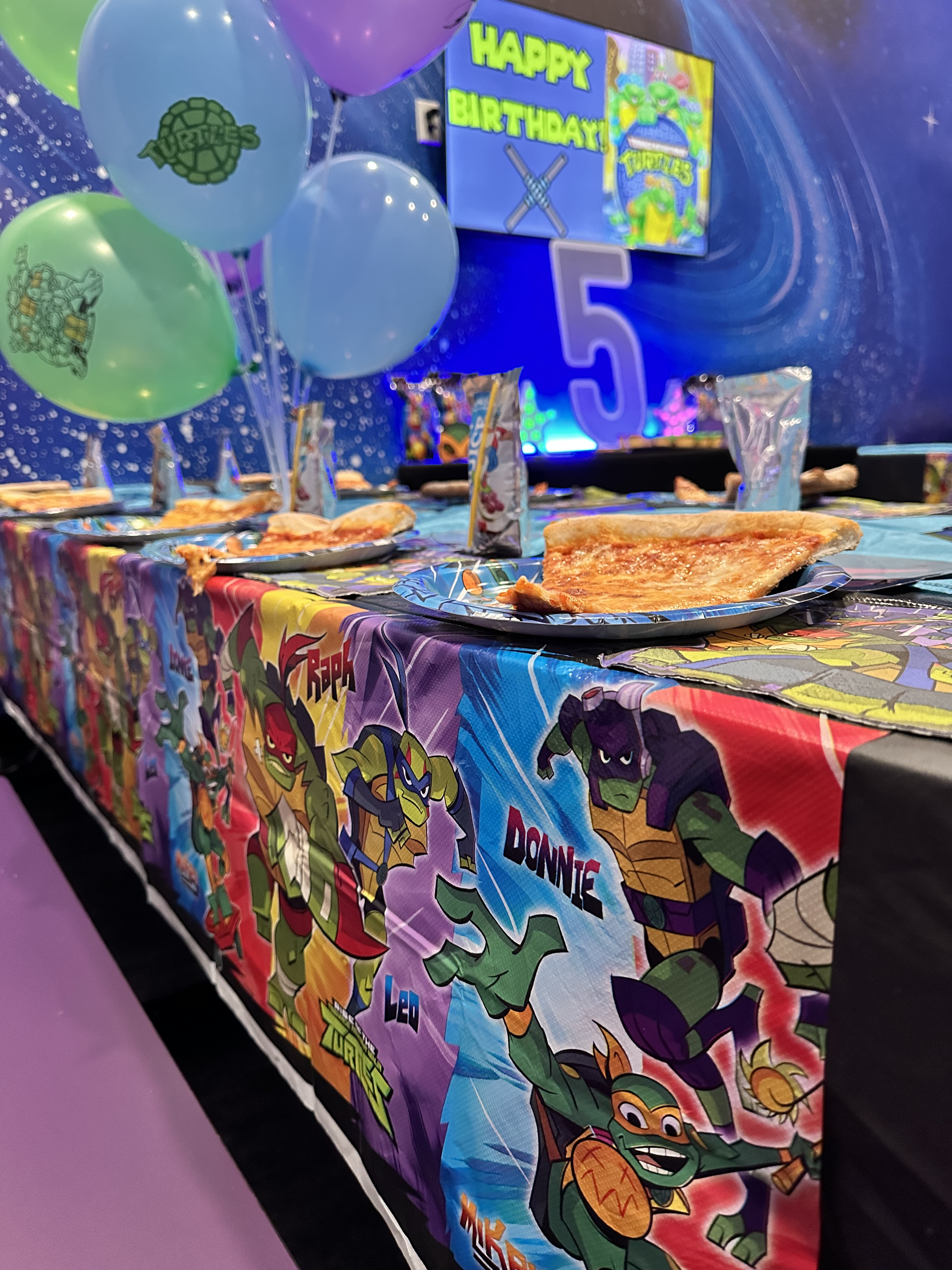 A colorful birthday party table decorated with Teenage Mutant Ninja Turtles themed tablecloth and balloons. There are balloons with Teenage Mutant Ninja Turtles logos, and a large digital screen displaying a "Happy Birthday" message with Teenage Mutant Ninja Turtles graphics. Several slices of pizza are on the table, along with party favor bags.