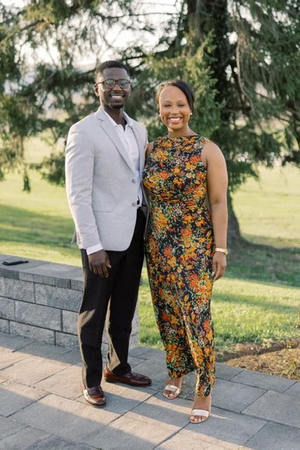 A smiling man and woman standing outdoors in front of trees, dressed in formal attire. The man wears a light gray suit jacket, white shirt, dark pants, and glasses. The woman wears a sleeveless floral dress and white heels.