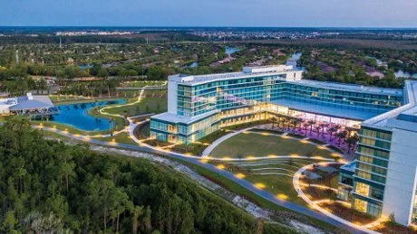 A modern multi-story office complex with glass facades, surrounded by landscaped gardens, walking paths, and a pond, set against a city skyline.