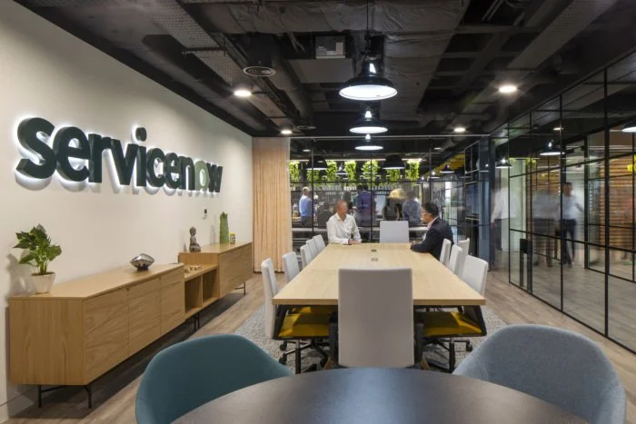 Modern office conference room with a long table, white chairs, and a glass wall. People are sitting and talking, with the company's logo 'ServiceNow' displayed on the wall.