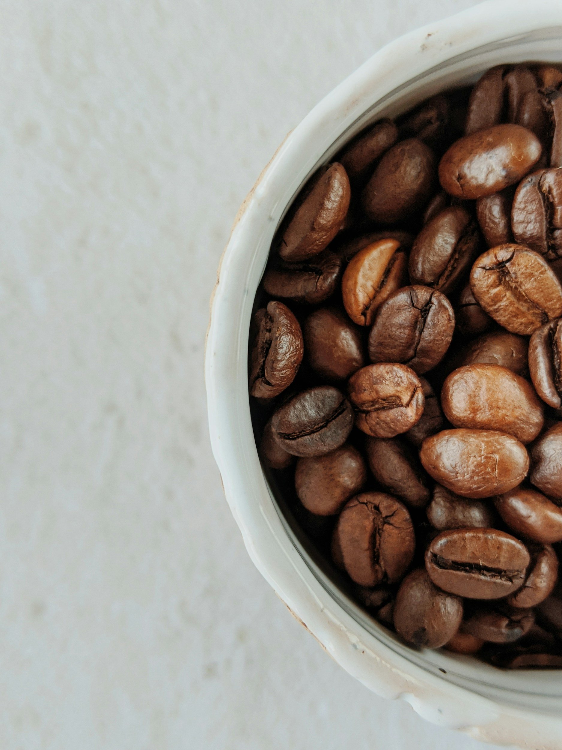 Close-up of a white cup filled with roasted seven-mile coffee beans on a light colored surface.