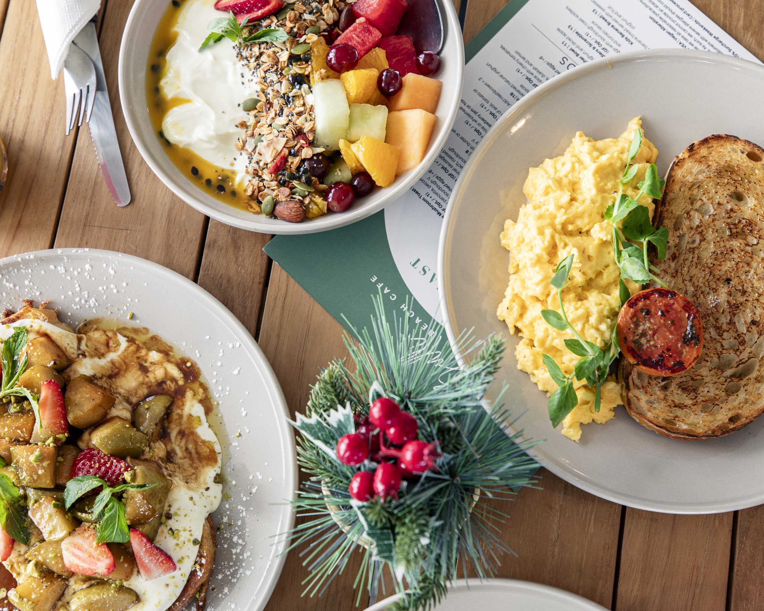 A top view of a breakfast table with plates of scrambled eggs with tomato, sourdough bread, and granola fruit bowl and fresh fruit and granola, along with a small holiday center piece and a breakfast menu and a French toast