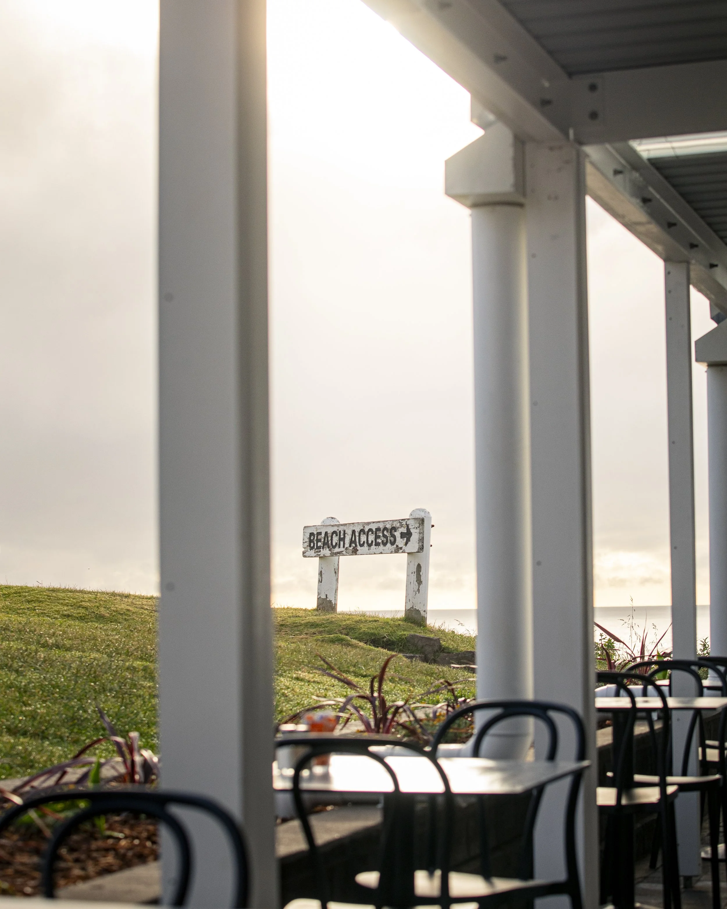 Outdoor view through a covered patio by the beach cafe with tables and chairs, showing a weathered sign reading 'Beach Access' with an arrow, on a hill leading to the beach at sunset or sunrise.