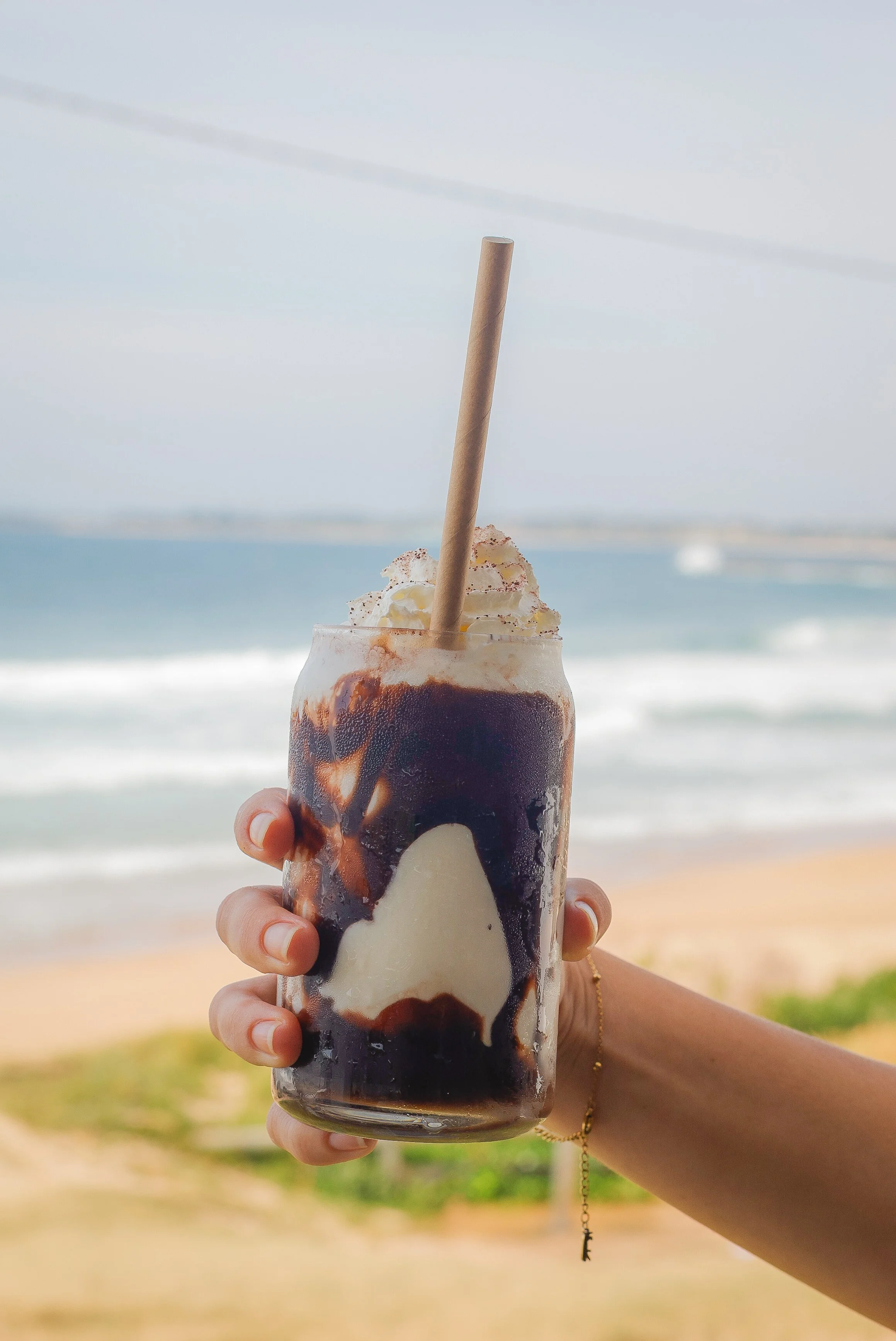 A hand holding a tall glass of a chocolate and vanilla ice cream float with whipped cream and a straw, set against a beach and ocean background.