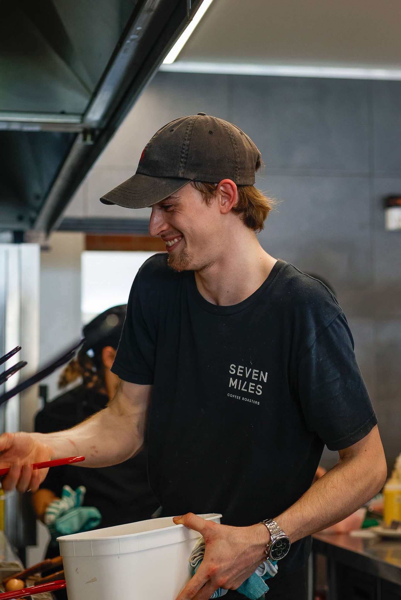 A young man wearing a black t-shirt with bakery text, a cap, and a watch, smiling while serving food at a cafe.