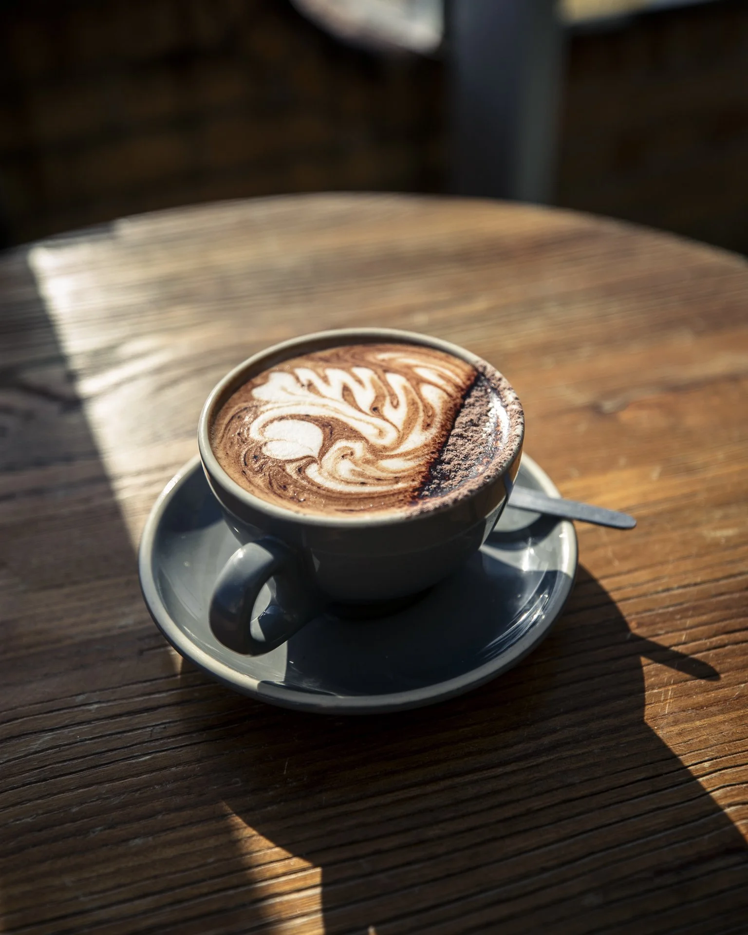 A cup of hot chocolate with foam art on top, sitting on a saucer with a spoon, placed on a wooden table with sunlight and shadow.