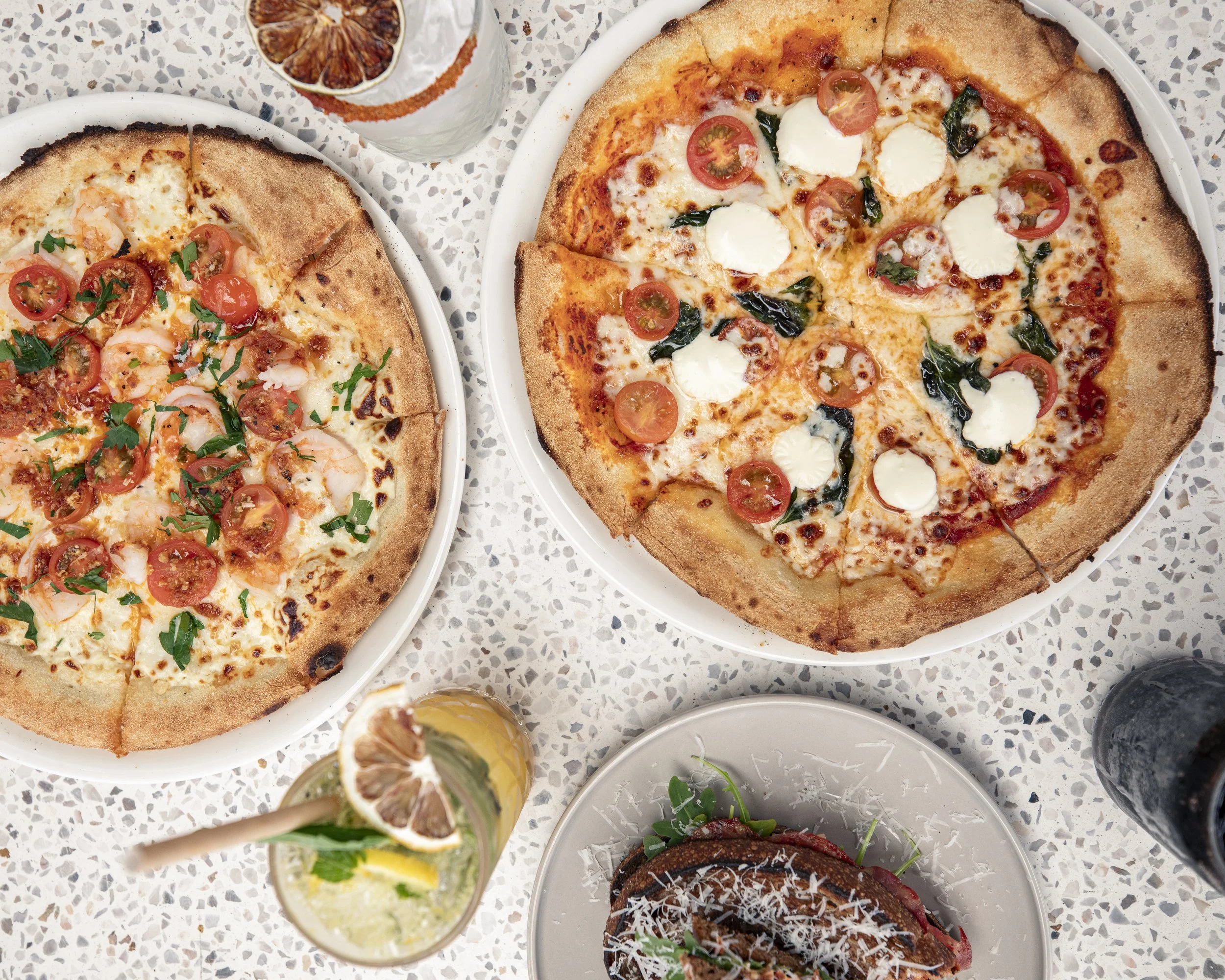 Two pizzas on white plates, one with cherry tomatoes, basil, and cheese, the other with cherry tomatoes, basil, and goat cheese; a cocktail with lemon and dried citrus slice; part of a sandwich with shredded cheese and greens; a dark bottle at the edge of the table.