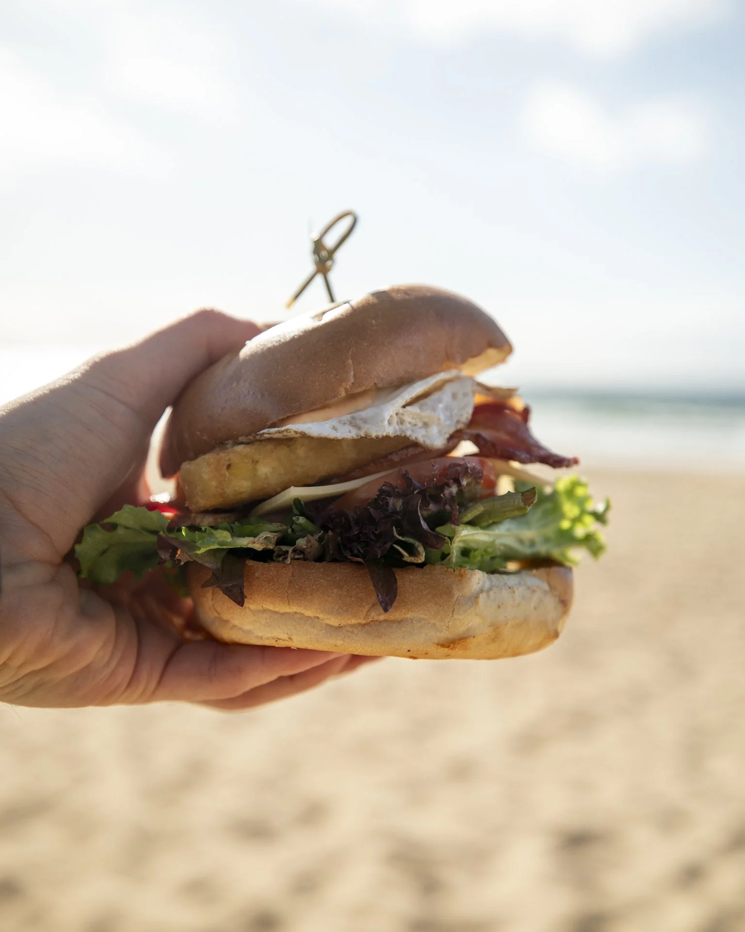 A person holding a sandwich with lettuce, bacon, egg, and sauce on a soft bun, against a beach background with sand and ocean.