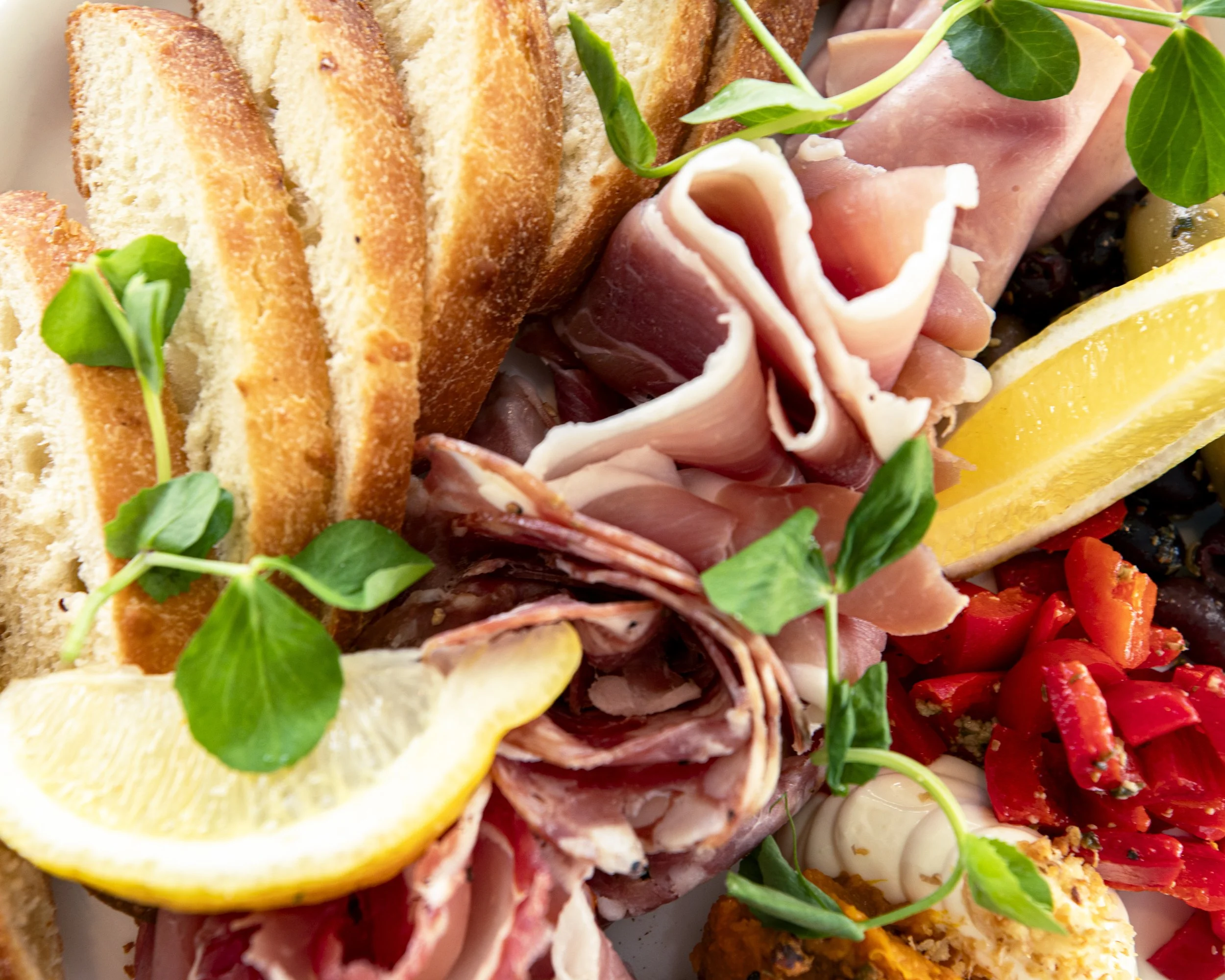 Close-up of a charcuterie platter with sliced bread, assorted cured meats, lemon wedges, red peppers, and green microgreens.