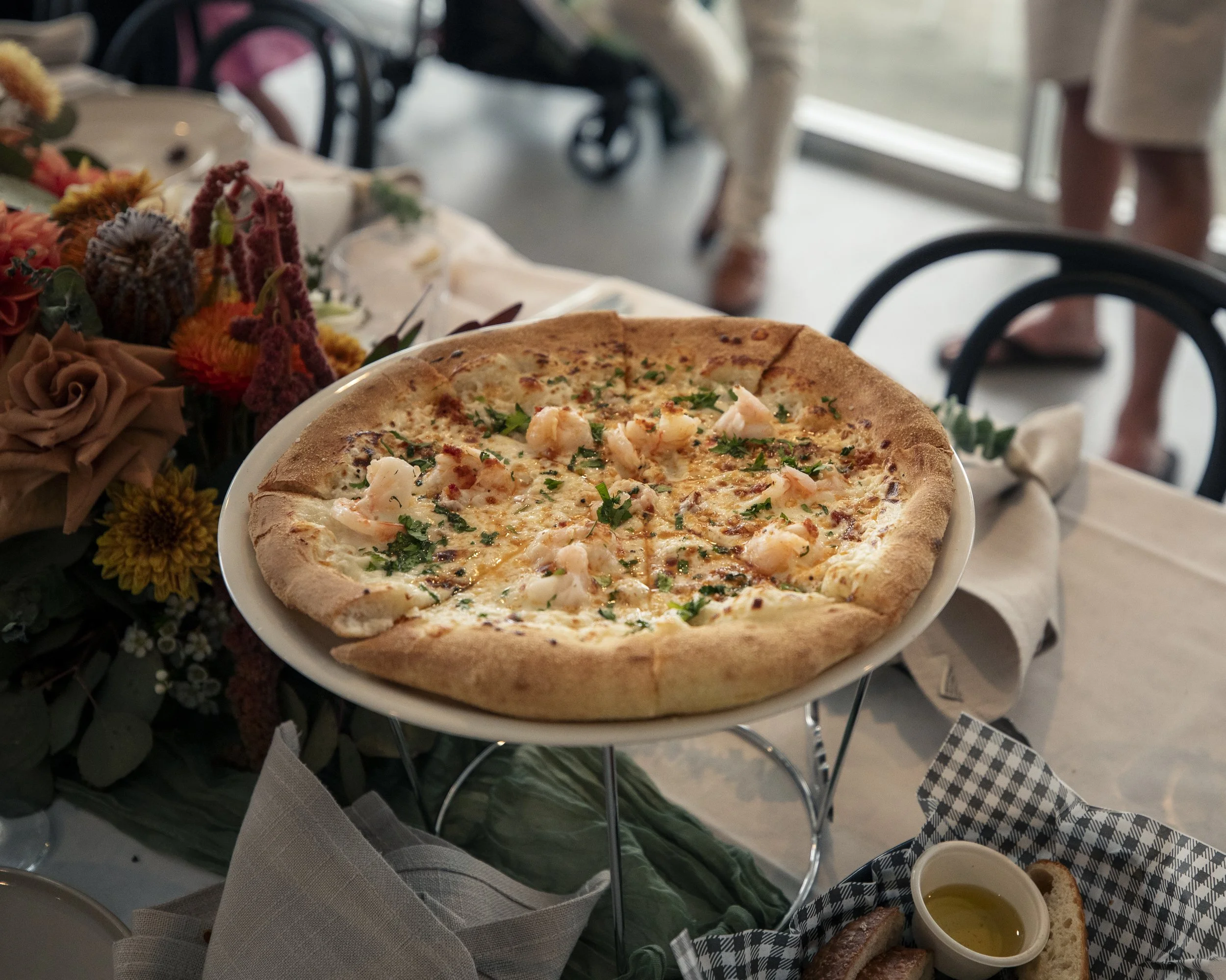 A prawn pizza on a white plate with chopped parsley, on a table with floral and linen decorations, and a side dish with oil and antipasto platter