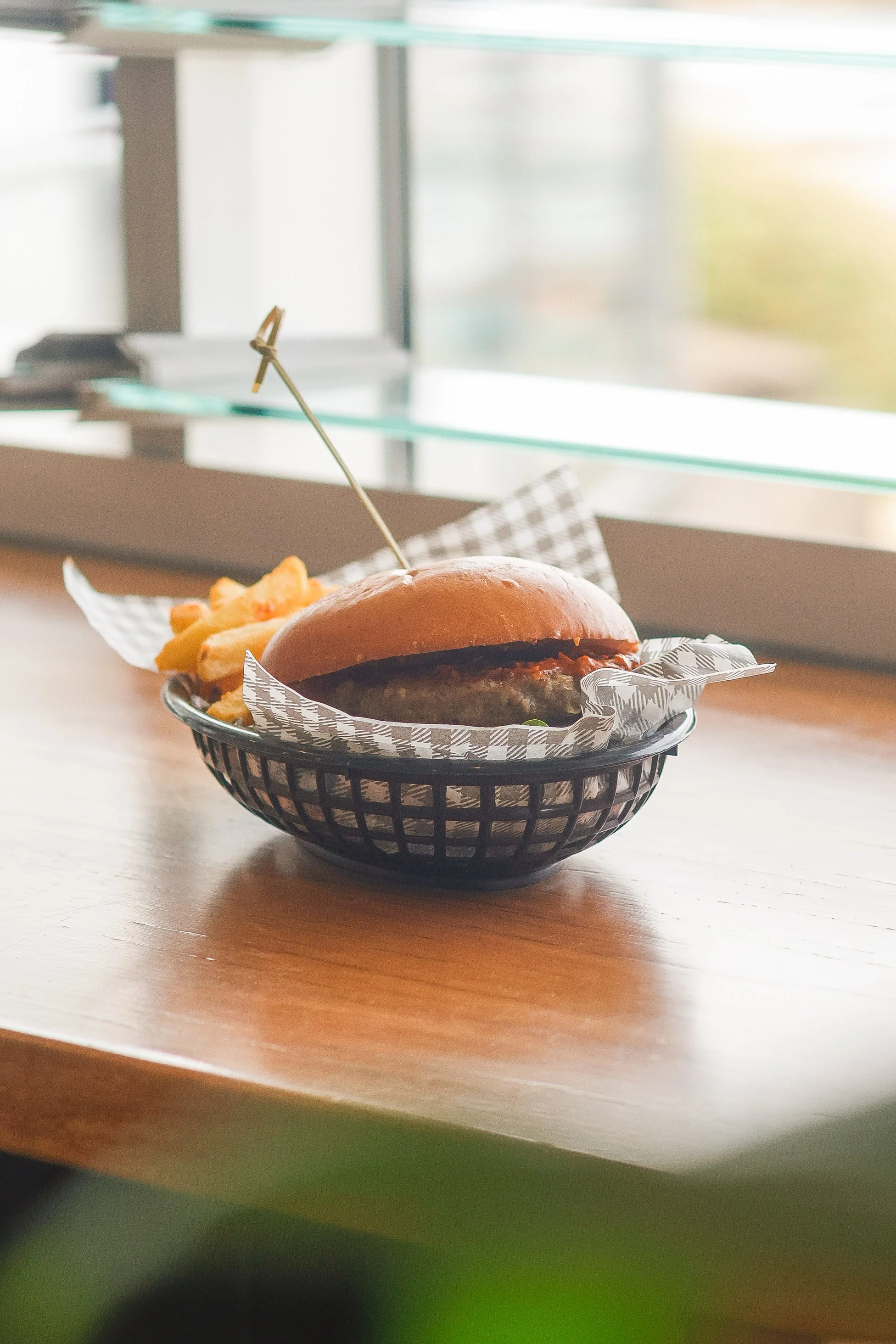 A burger with fries in a black basket on a wooden table near a window, sunlight streaming in.