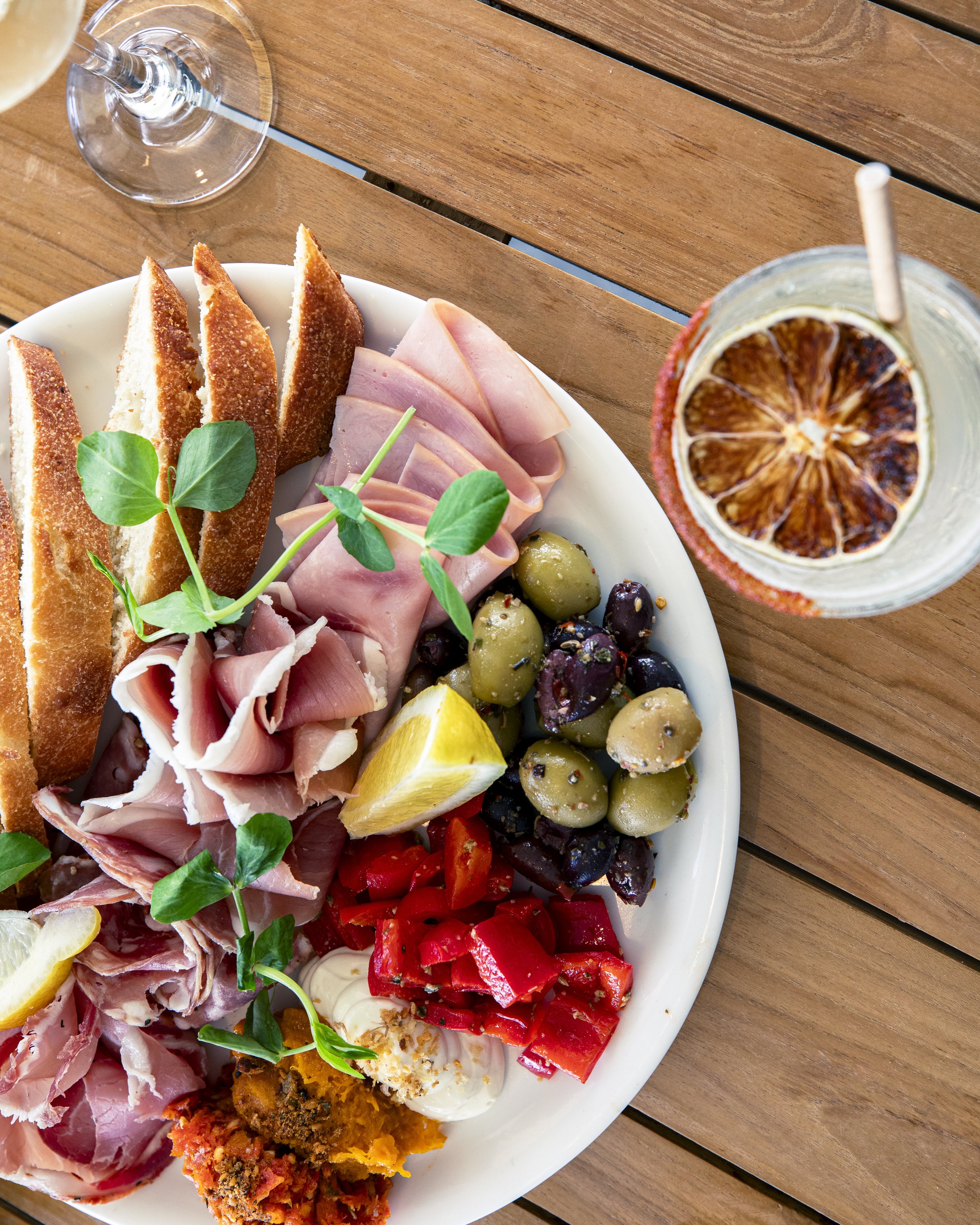 Charcuterie board with sliced bread, ham, prosciutto, olives, red peppers, lemon wedges, and garnished with microgreens on a wooden table, with a glass of wine and a drink with a dried lemon slice.