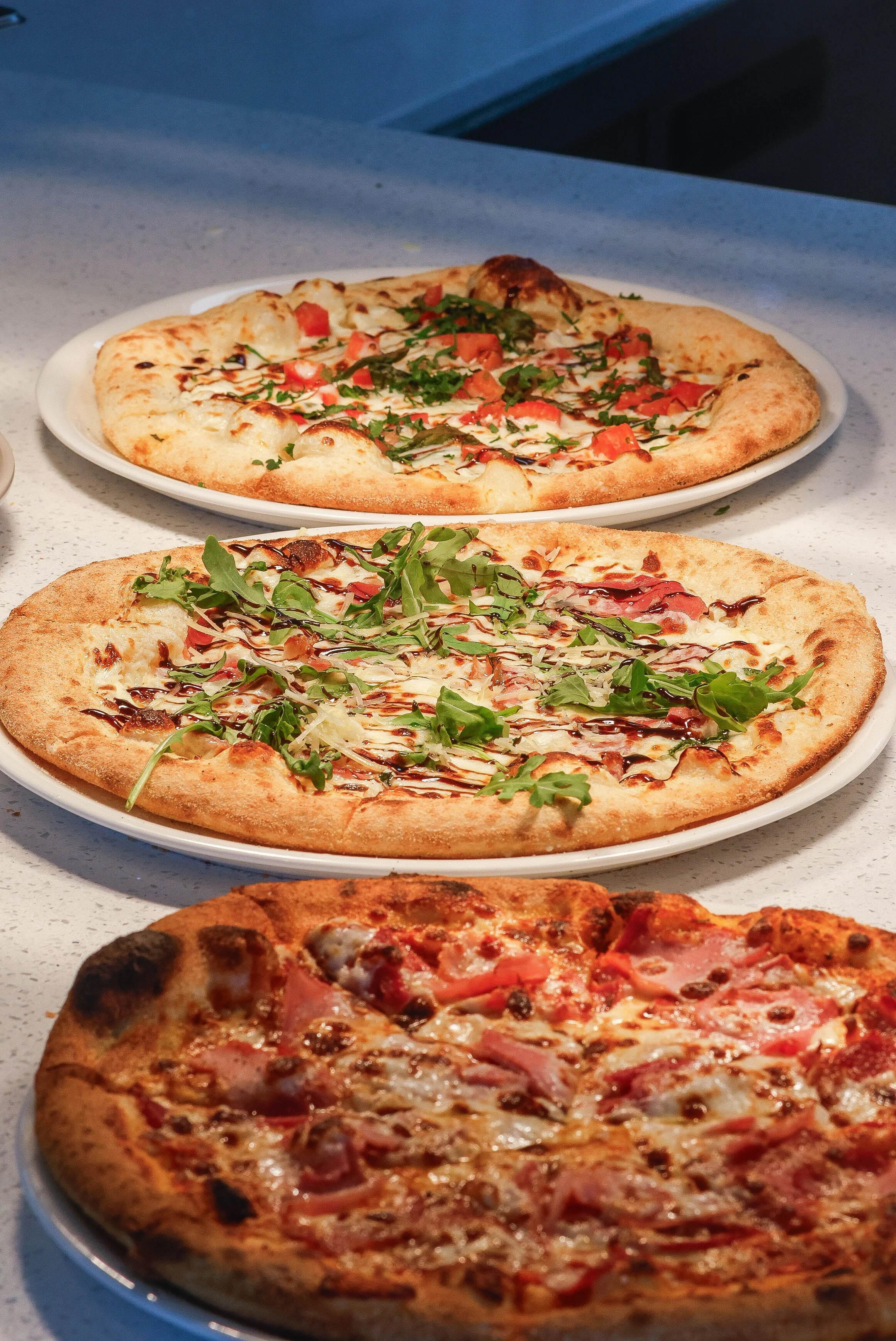 Three plates of wood fired pizza on a white marble table, the pizzas are a wood fired meat lovers pizza, bruschetta pizza, and a prawn pizza.