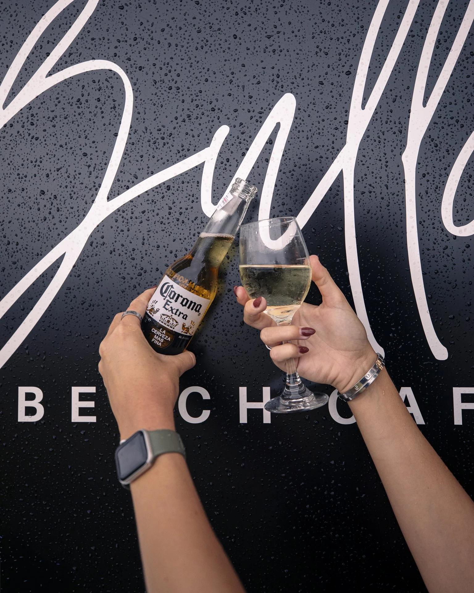 Two people toasting with a bottle of Corona beer and a glass of white wine in front of a black background with rain droplets and white stylized text.