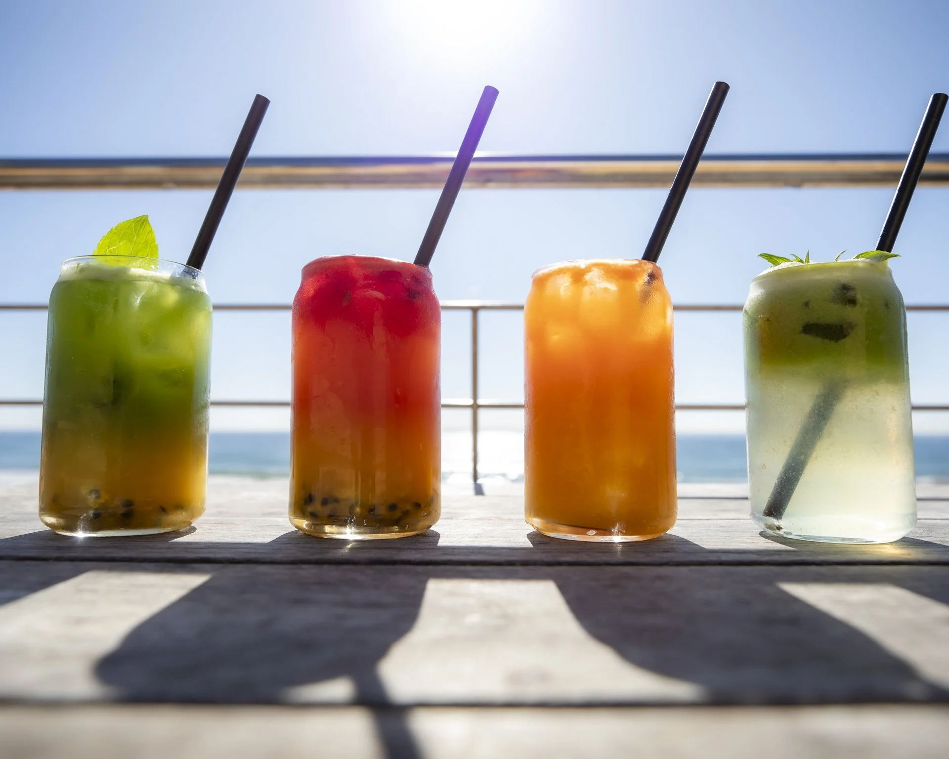 Four colorful fruit juices with black straws on a wooden surface outdoors with a bright blue sky and ocean in the background.