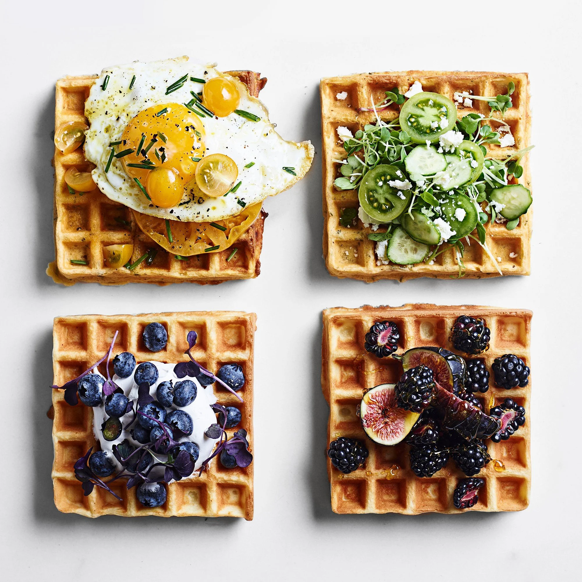 Four square waffles topped with various toppings on a white background, including fried eggs and greens, blueberries and whipped cream, and blackberries with figs.