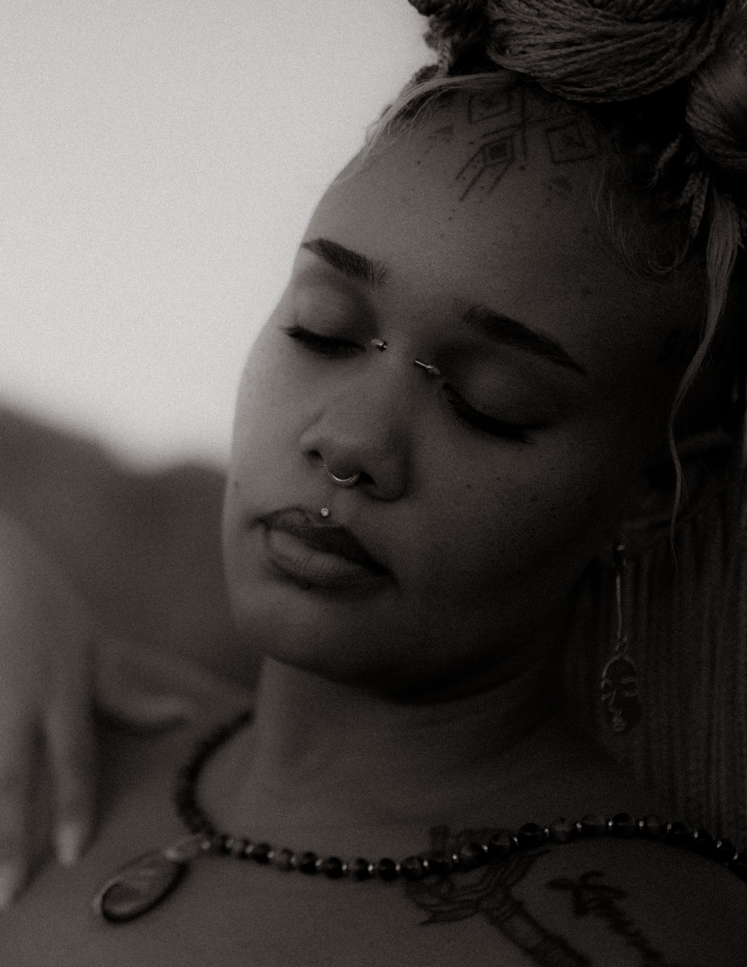 A young woman with braided hair, multiple facial piercings, and tattoos, wearing jewelry, with her eyes closed.