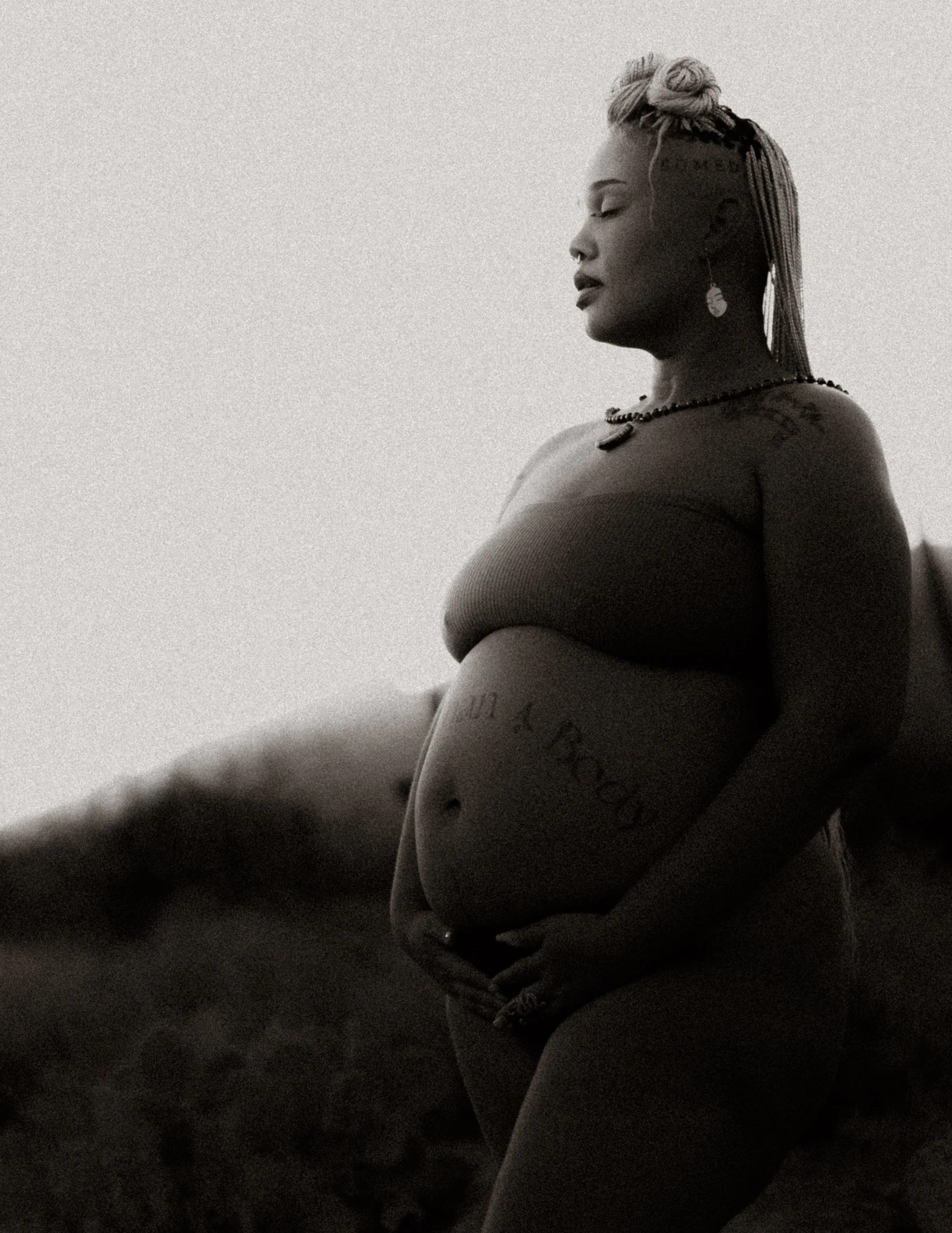 A side profile of a pregnant woman standing outdoors with eyes closed. She wears a tube top, jewelry, and has a hairstyle with braided design on the side and buns on top. The background appears to be natural landscape or desert.